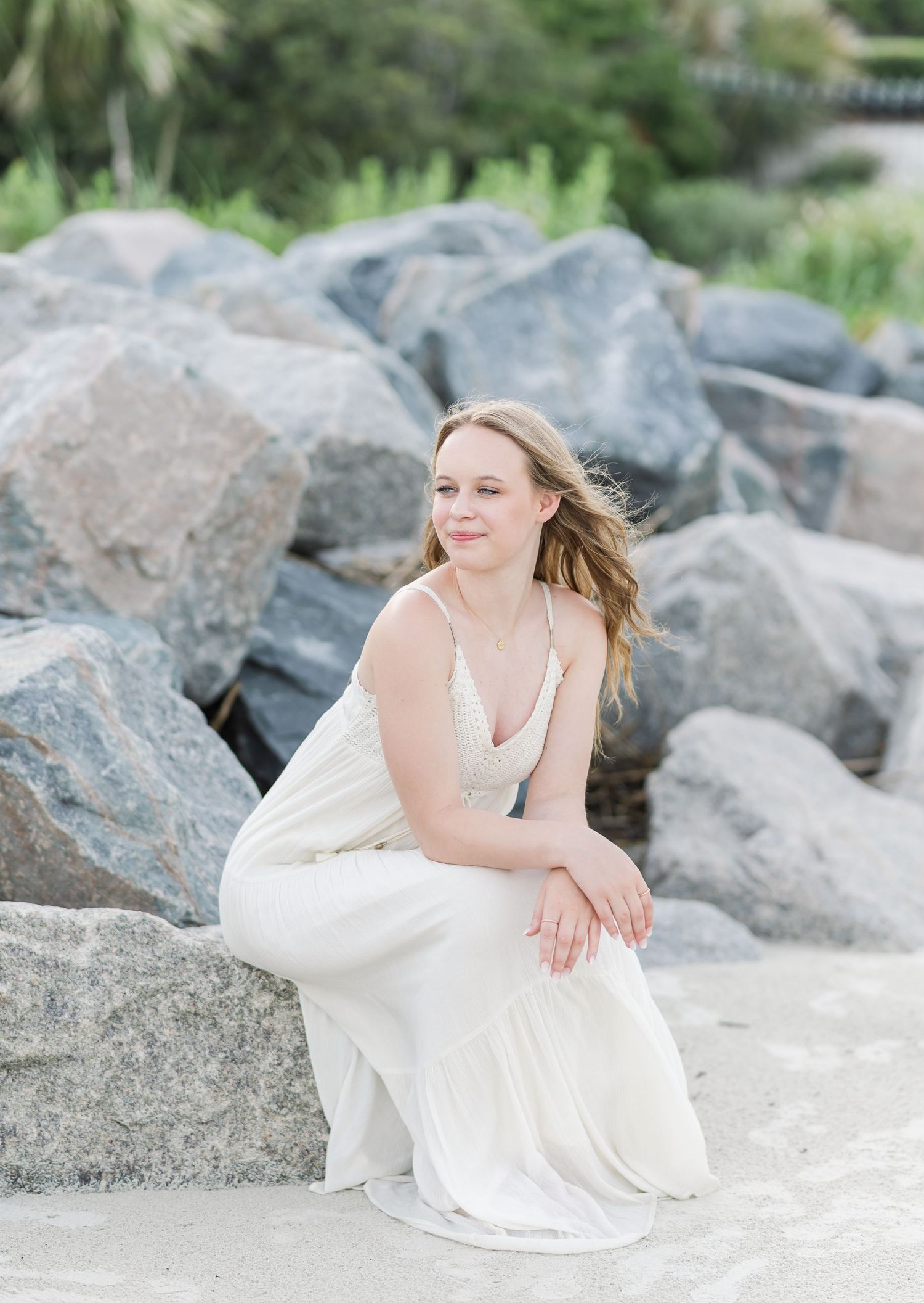 A person in a white dress sits gracefully on coastal rocks in an elegant outdoor portrait.