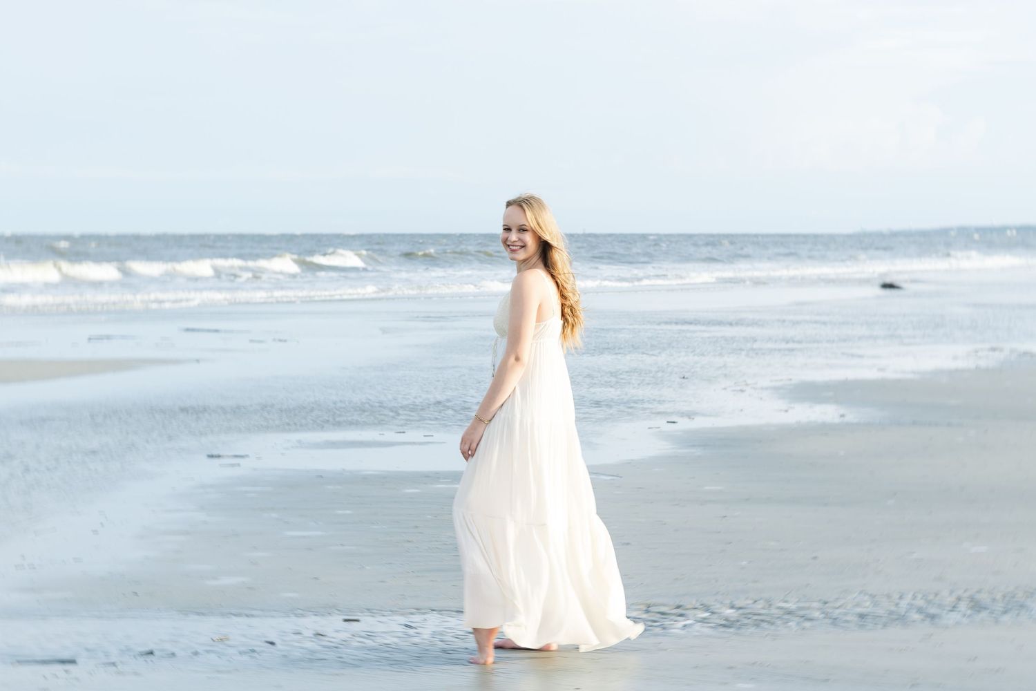 A graceful figure in a flowing white dress walks barefoot along a serene beach shoreline on an overcast day.