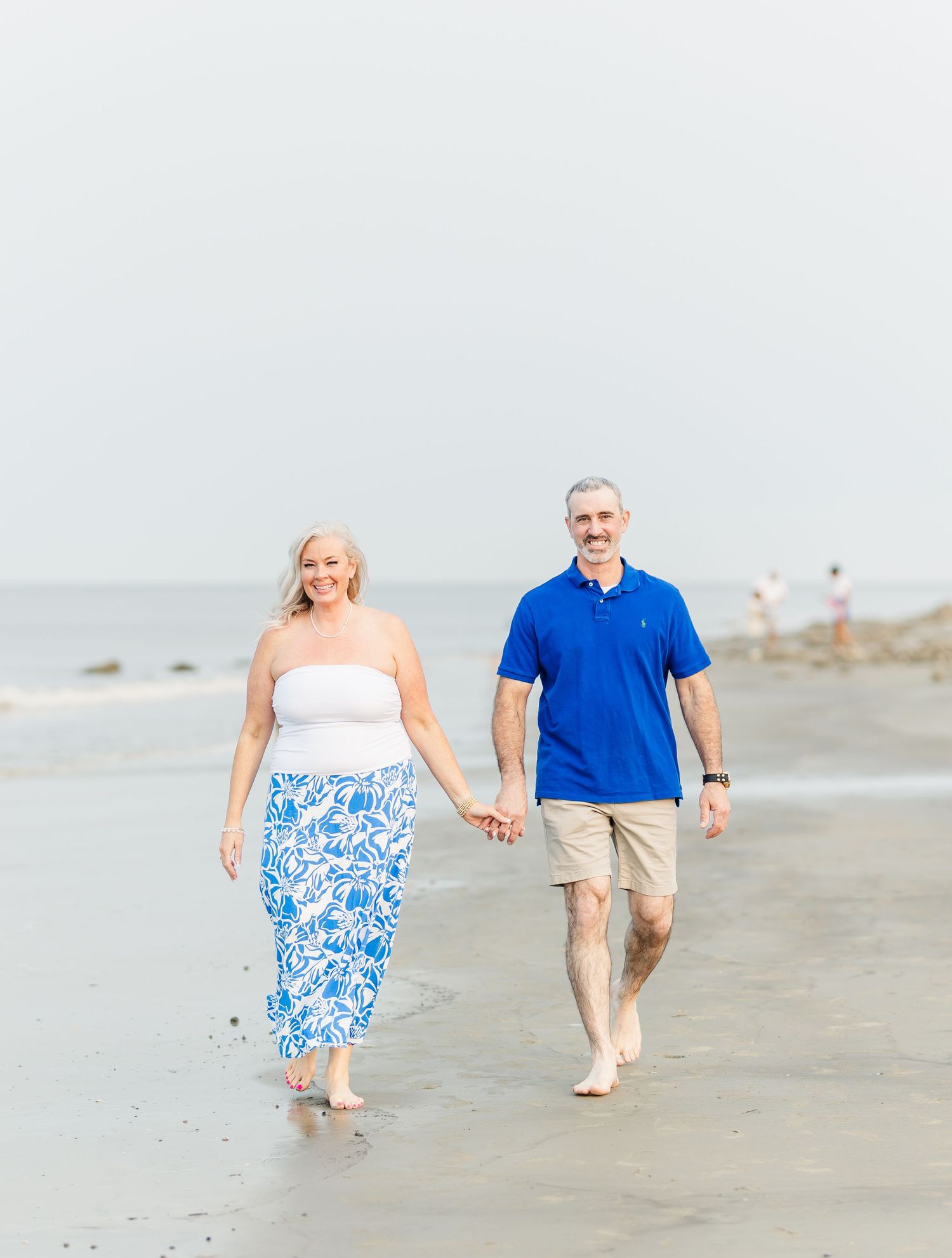 A couple walks hand in hand along a beach wearing coordinating blue and white outfits in a romantic seaside scene.