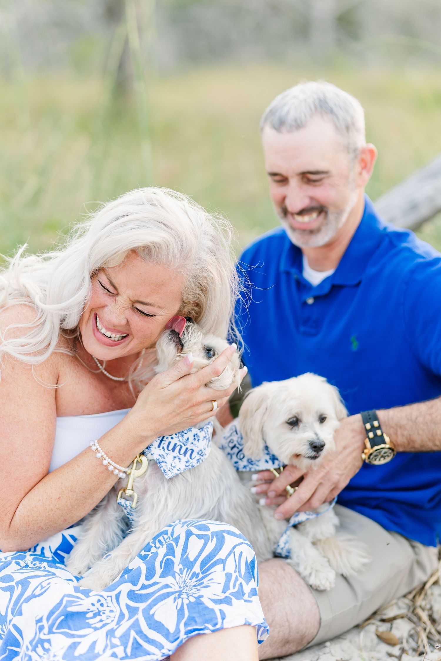 A couple sits in a grassy field playing with two small white dogs while wearing coordinated blue and white outfits.