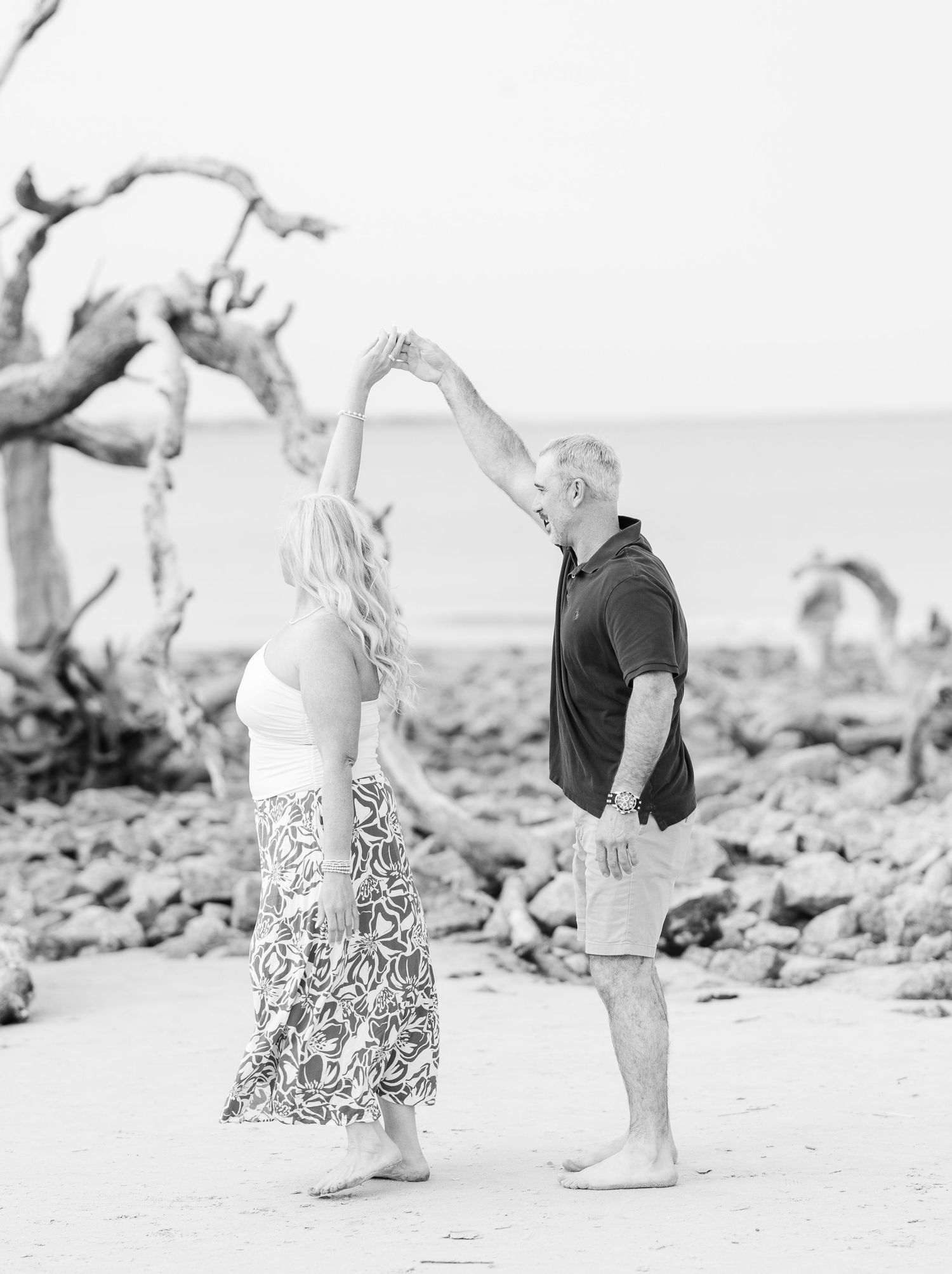 A romantic black and white photo of a couple dancing together on a driftwood-lined beach.