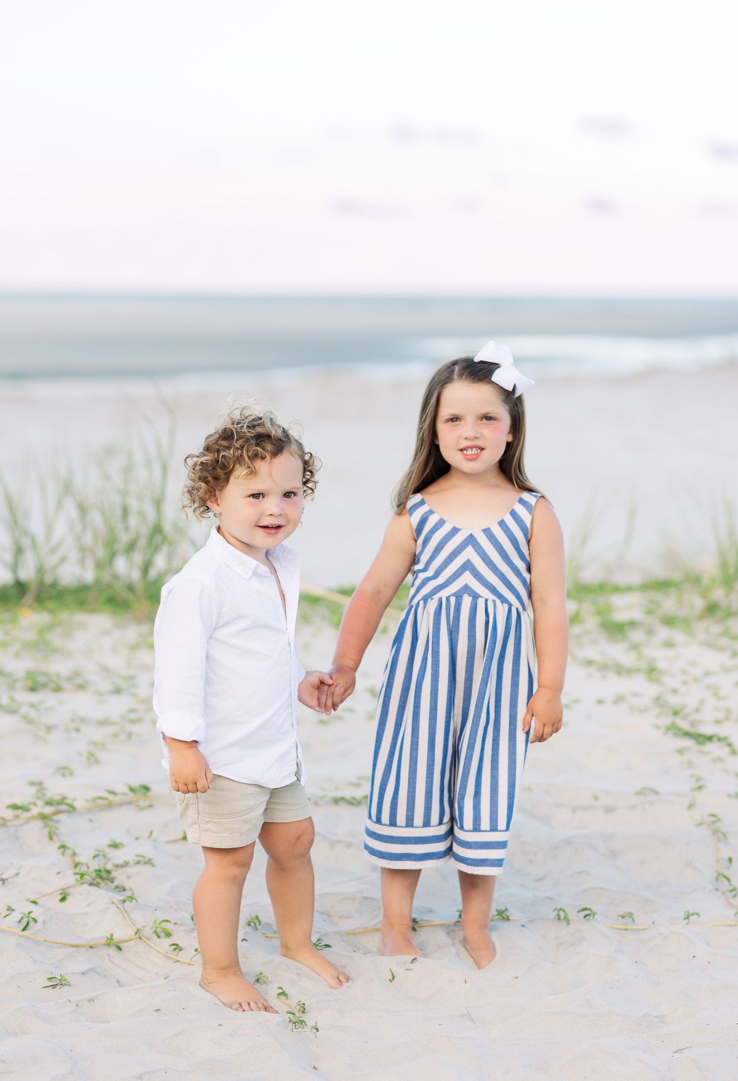 Two children wearing summer outfits play together on a sandy beach with sparse vegetation.