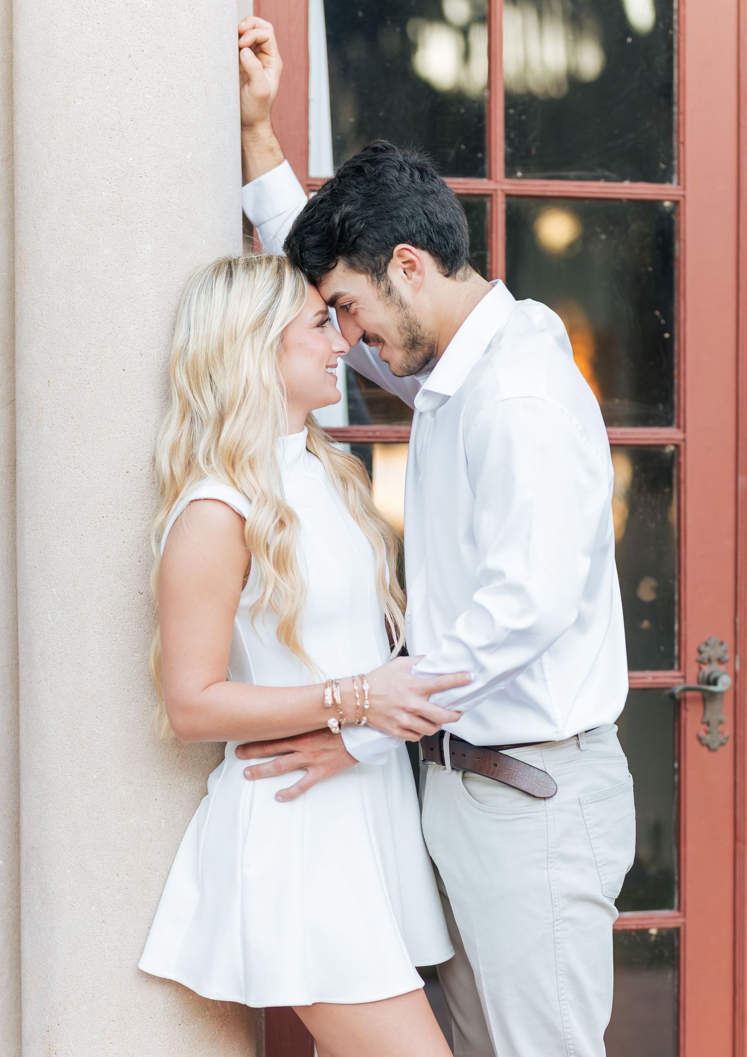 A couple in white outfits share a romantic moment leaning against a column near a red door.