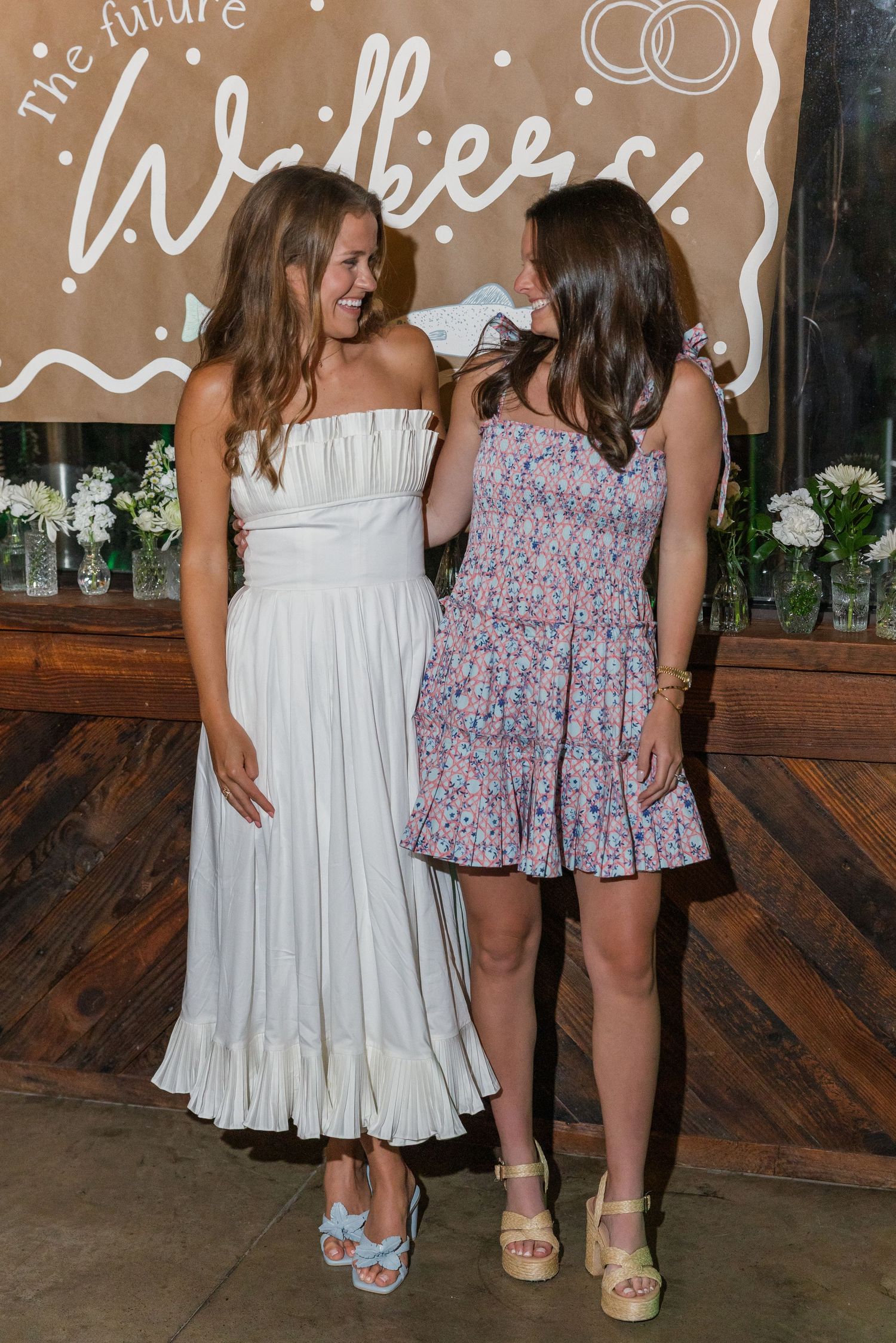 Bride-to-be in white dress laughs with friend in floral mini dress at engagement party.