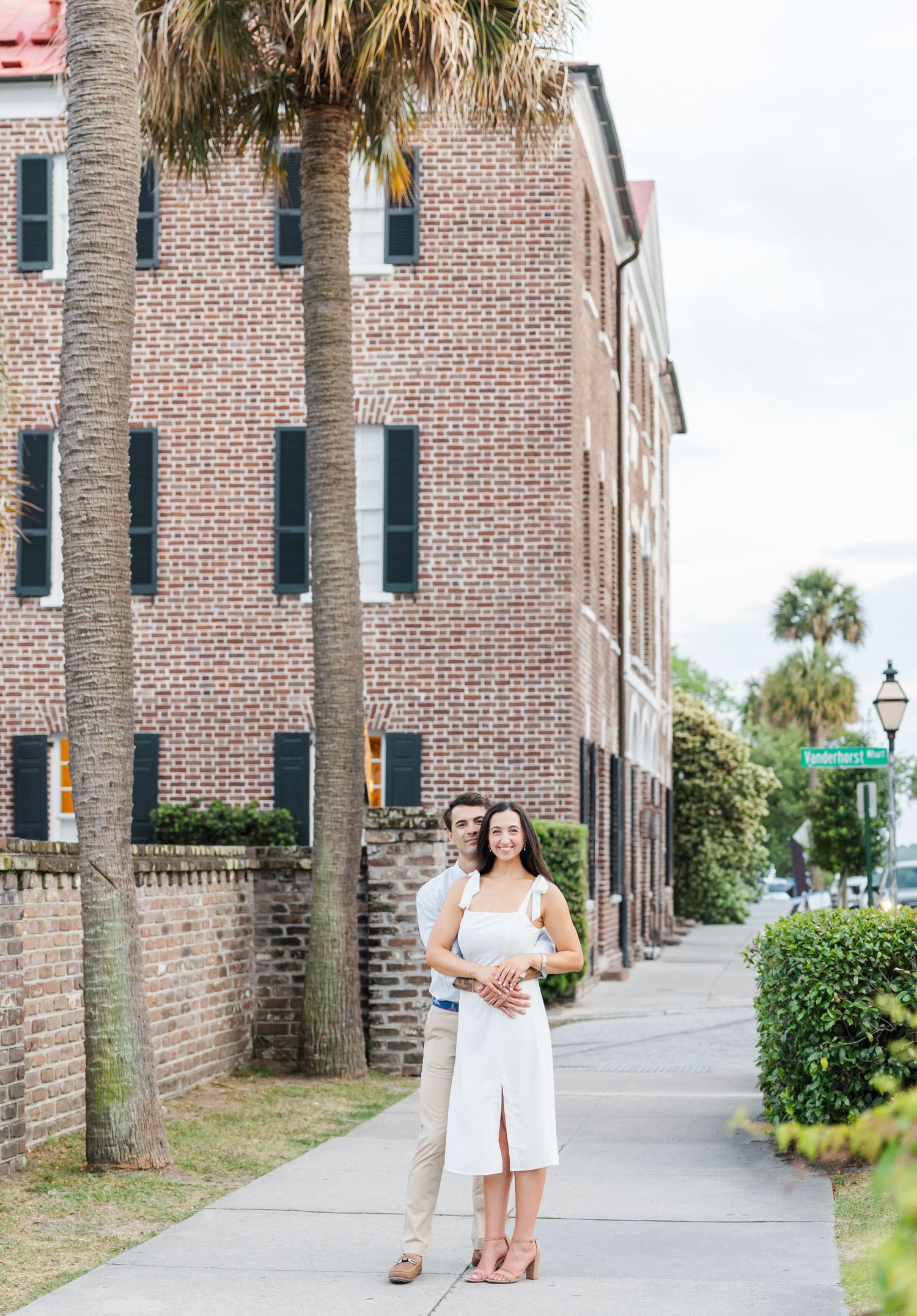Engaged couple embracing on a Charleston sidewalk, woman in white dress, man in white shirt, historic brick buildings behind.