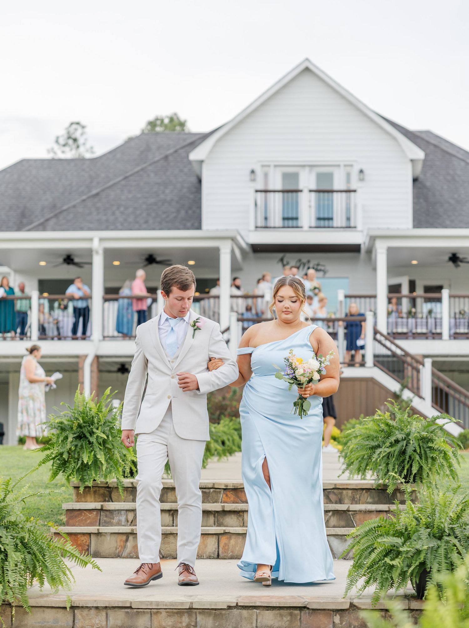 Groomsman in beige suit escorts bridesmaid in light blue one-shoulder gown down wedding ceremony steps.