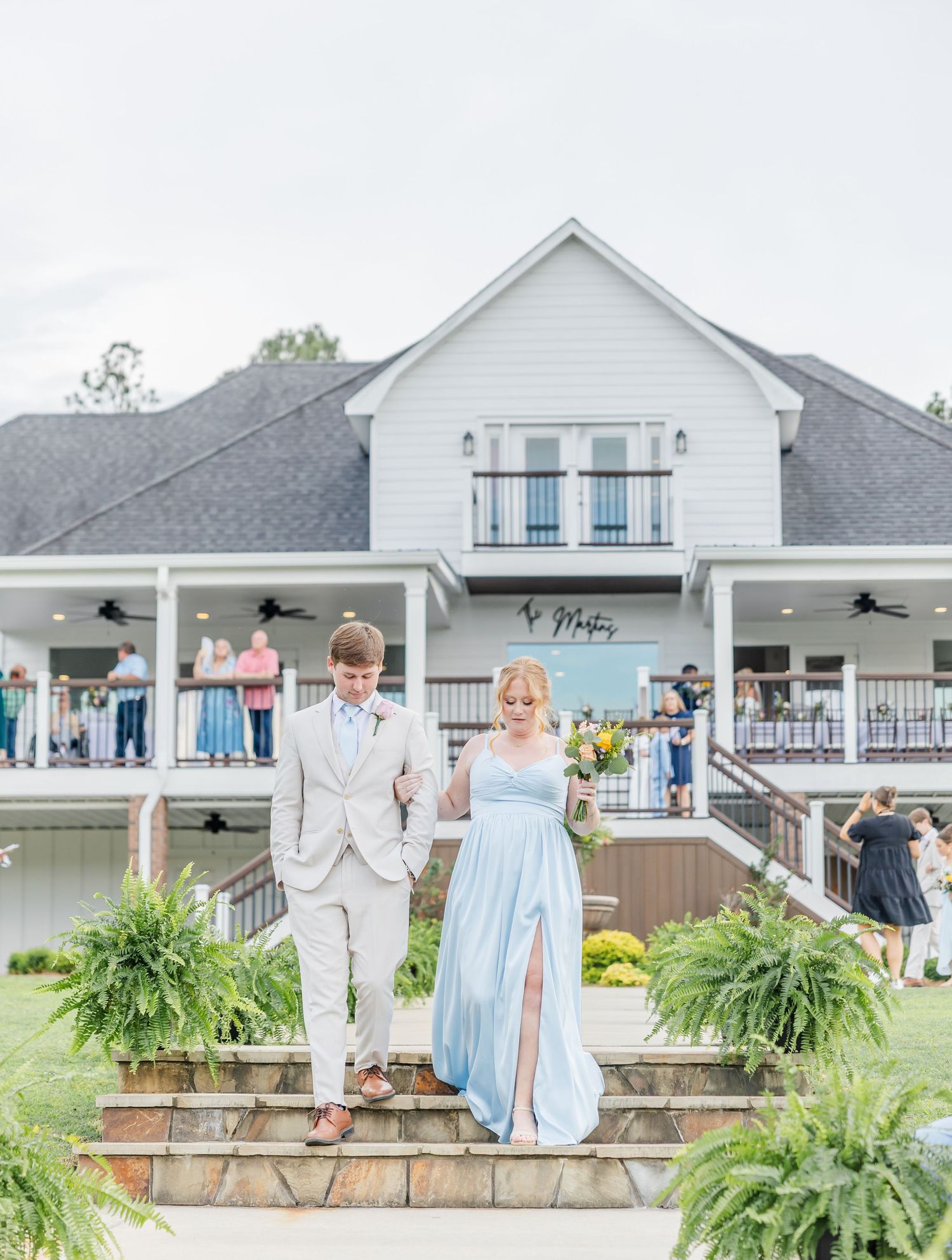 A bridesmaid in a blue gown and groomsman in a tan suit walk hand-in-hand down steps at a wedding venue.