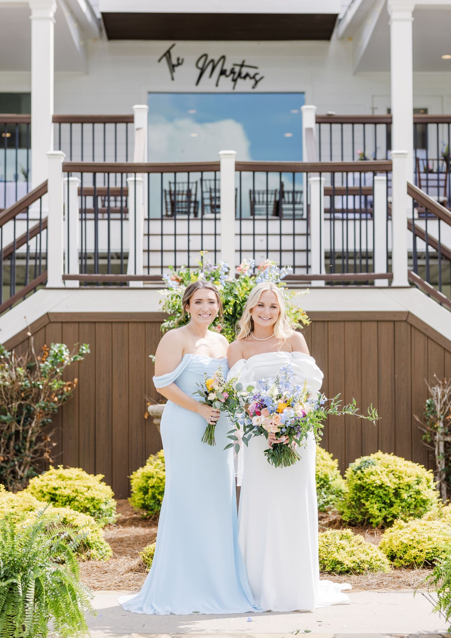 Two women in white and light blue gowns holding bouquet outside wedding venue.