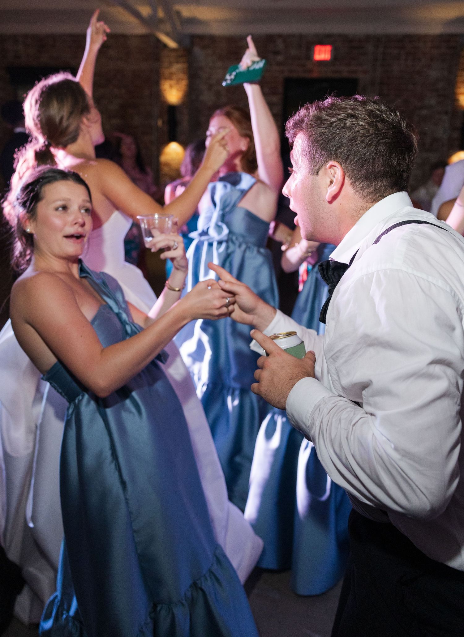Bridesmaid in blue gown and groomsman in suspenders dance together at vibrant wedding reception.