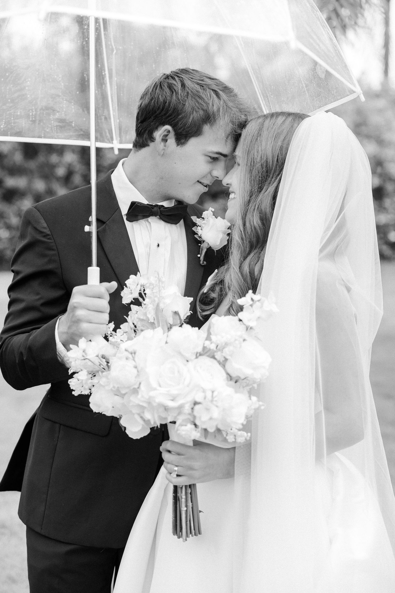 Black and white photo of bride and groom sharing romantic moment under clear umbrella with white rose bouquet.