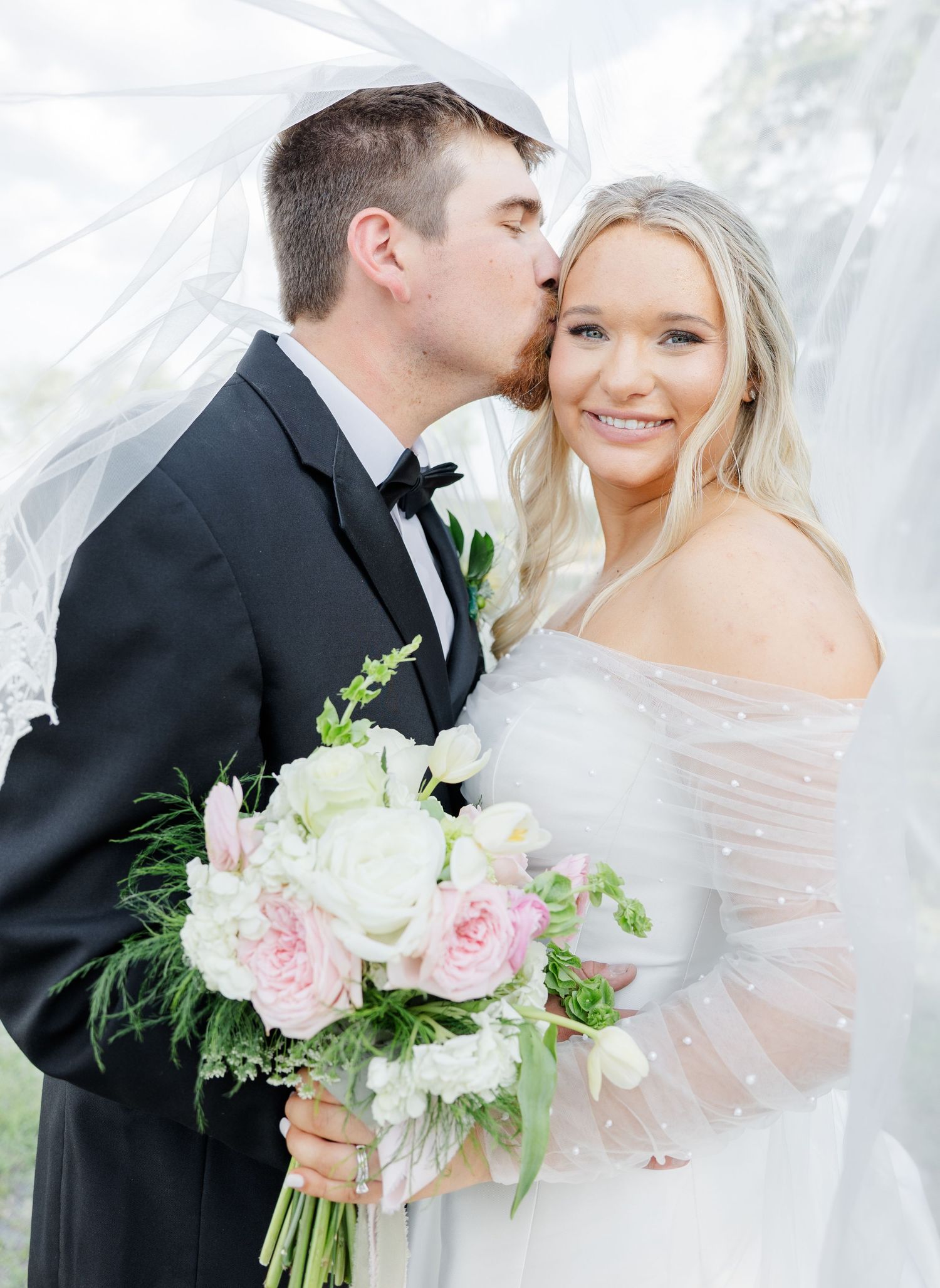 Groom in black tuxedo kissing bride on cheek while she smiles holding pink and white bouquet.