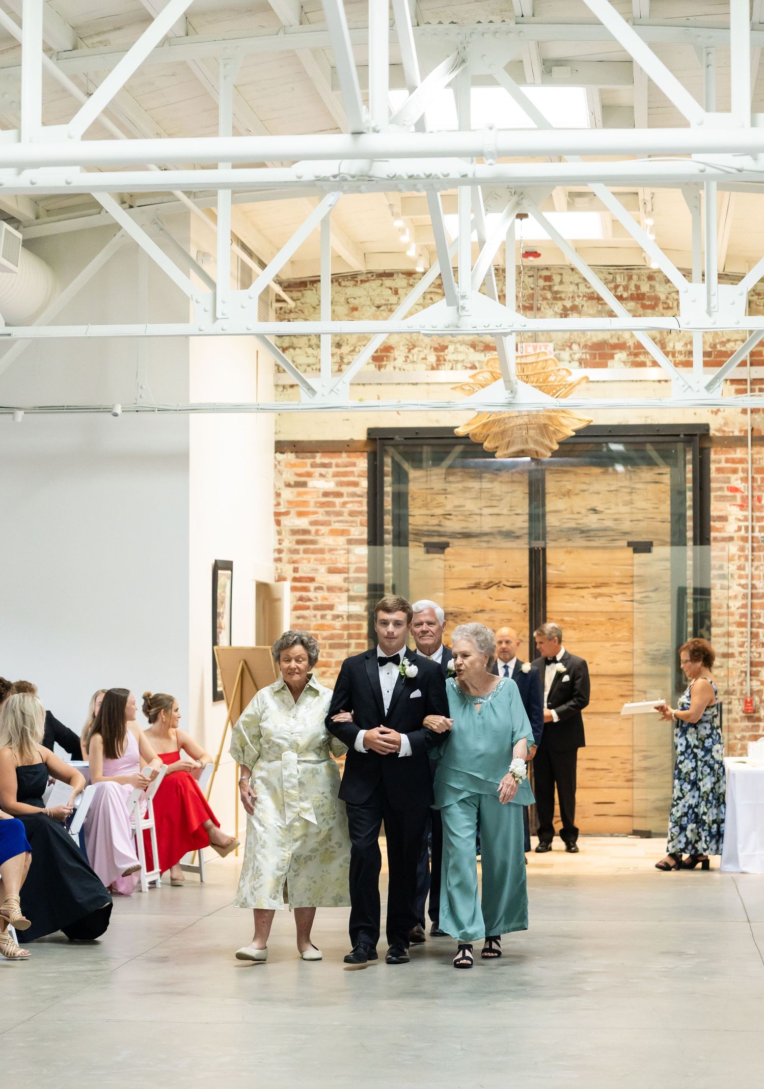 Groom escorts two elegantly dressed mothers down the aisle at an industrial chic wedding venue with exposed brick.