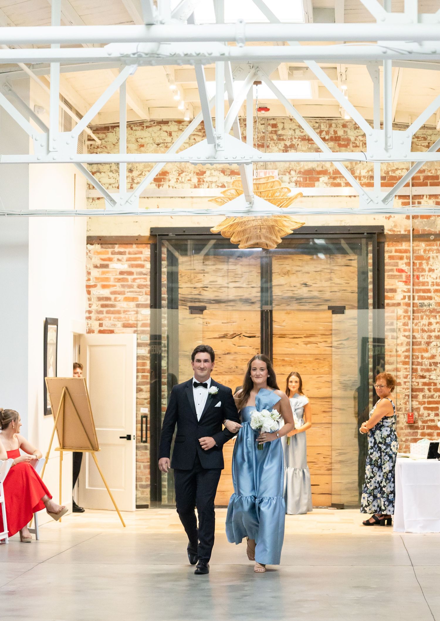 Groomsman escorts bridesmaid in blue dress down aisle at modern industrial wedding venue.