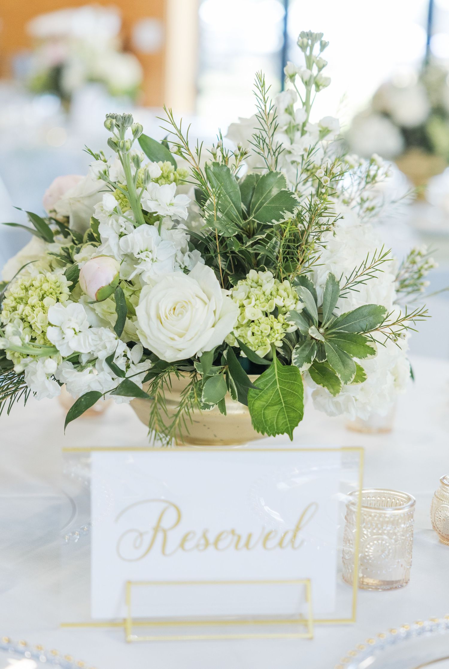 White and green floral wedding centerpiece with roses and hydrangeas beside a gold-framed Reserved table sign.