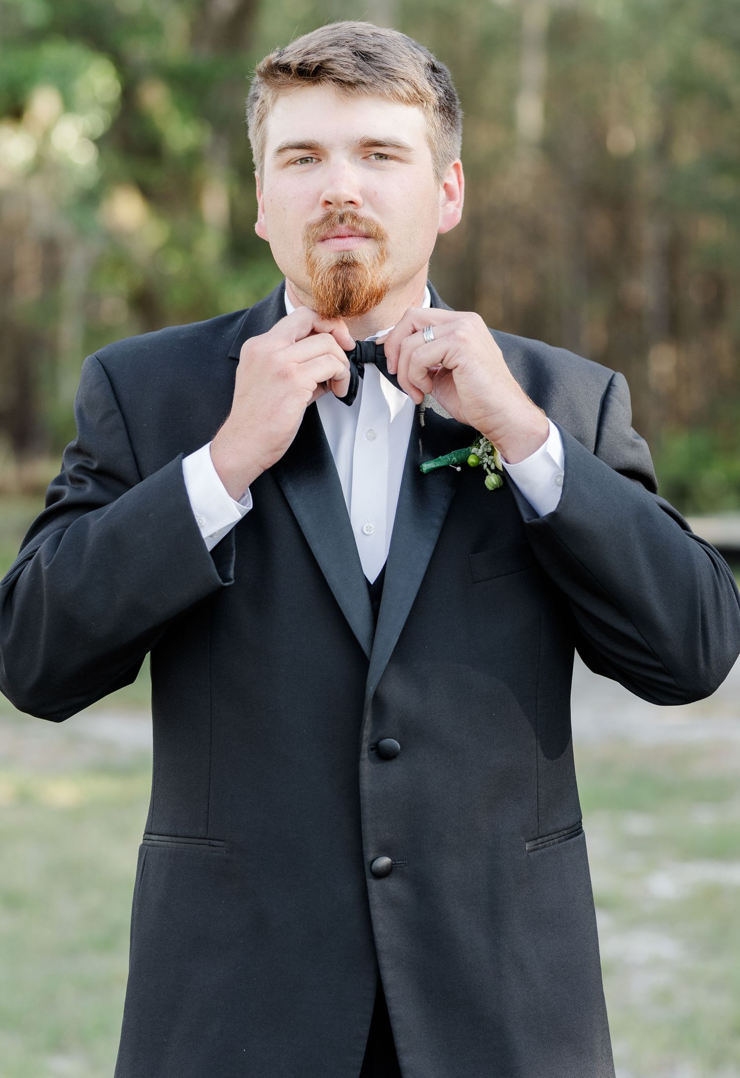 Groom in black tuxedo adjusting bow tie with green boutonniere outdoors.