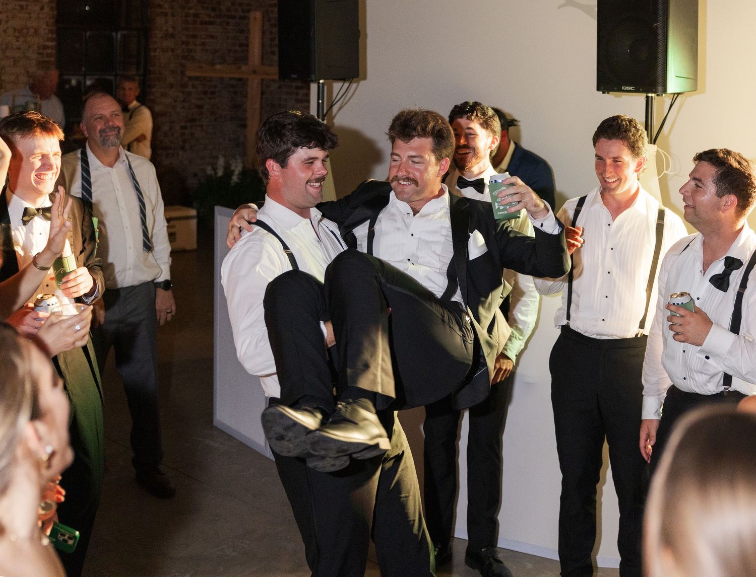 Groomsmen in tuxedos and suspenders lift and celebrate with groom on the dance floor at reception.
