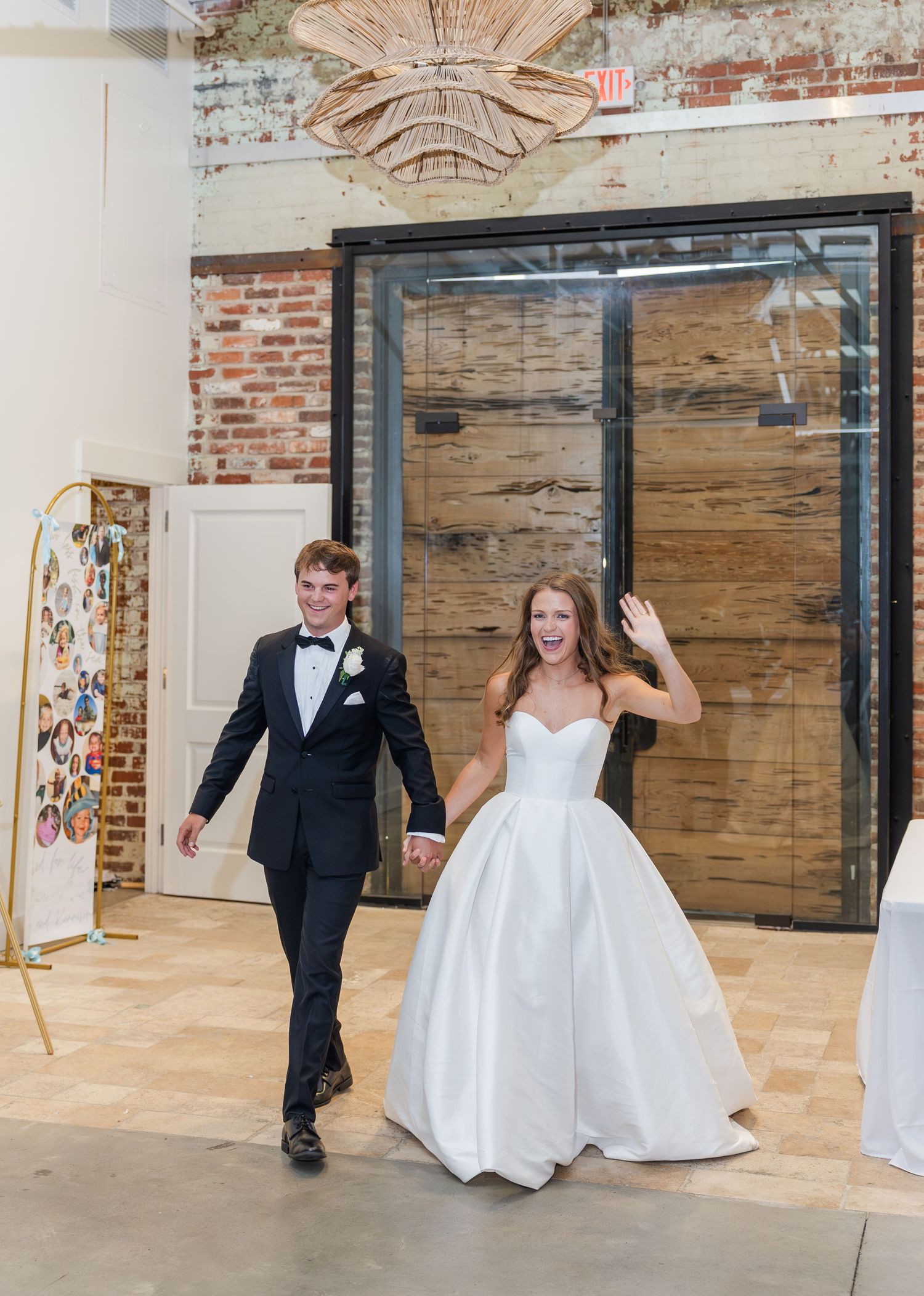 Bride in ballgown waving as she enters reception hall with groom in black tuxedo through wooden doors.