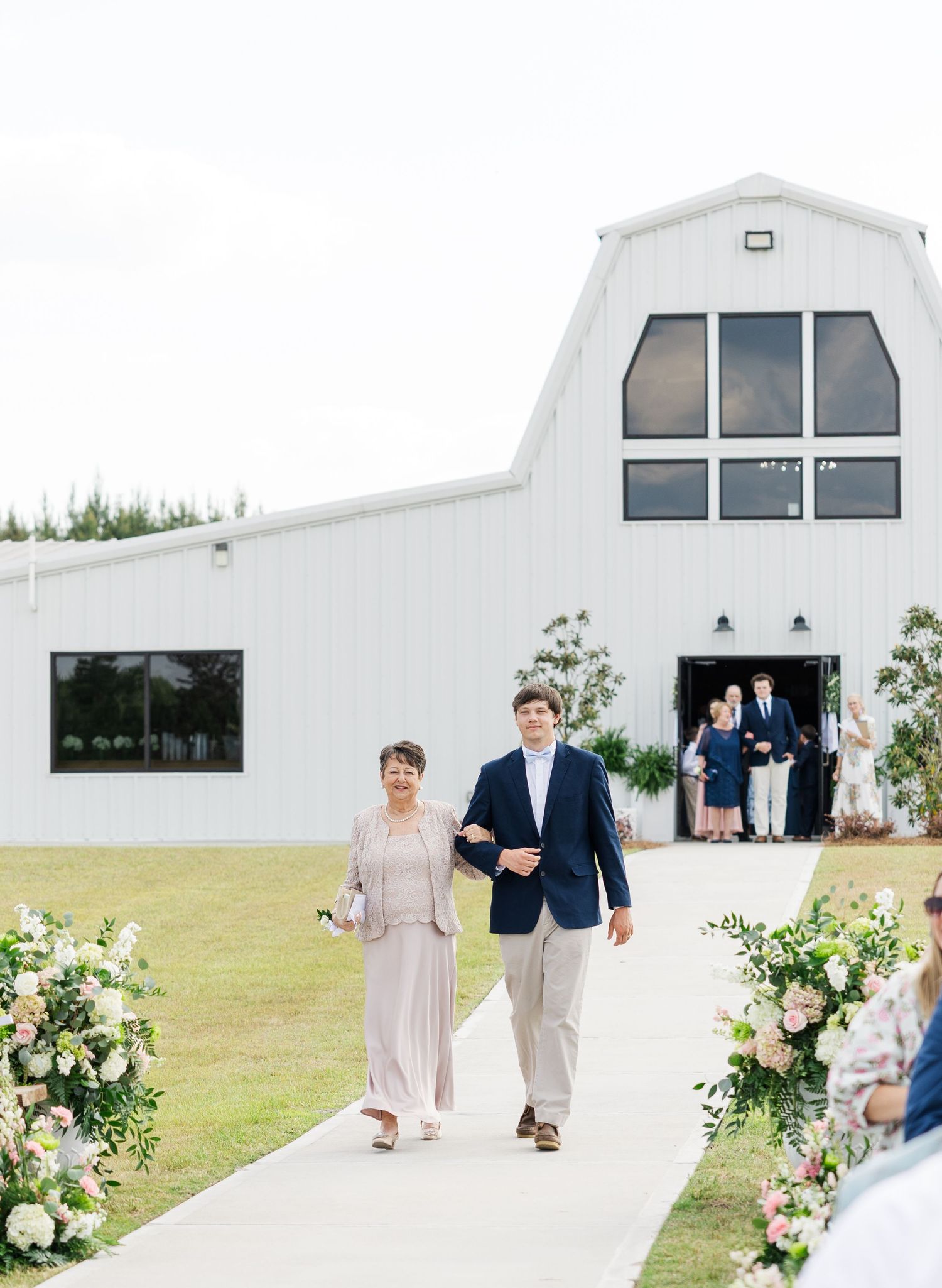 Groomsman escorts woman down floral-lined outdoor aisle toward white barn wedding venue.