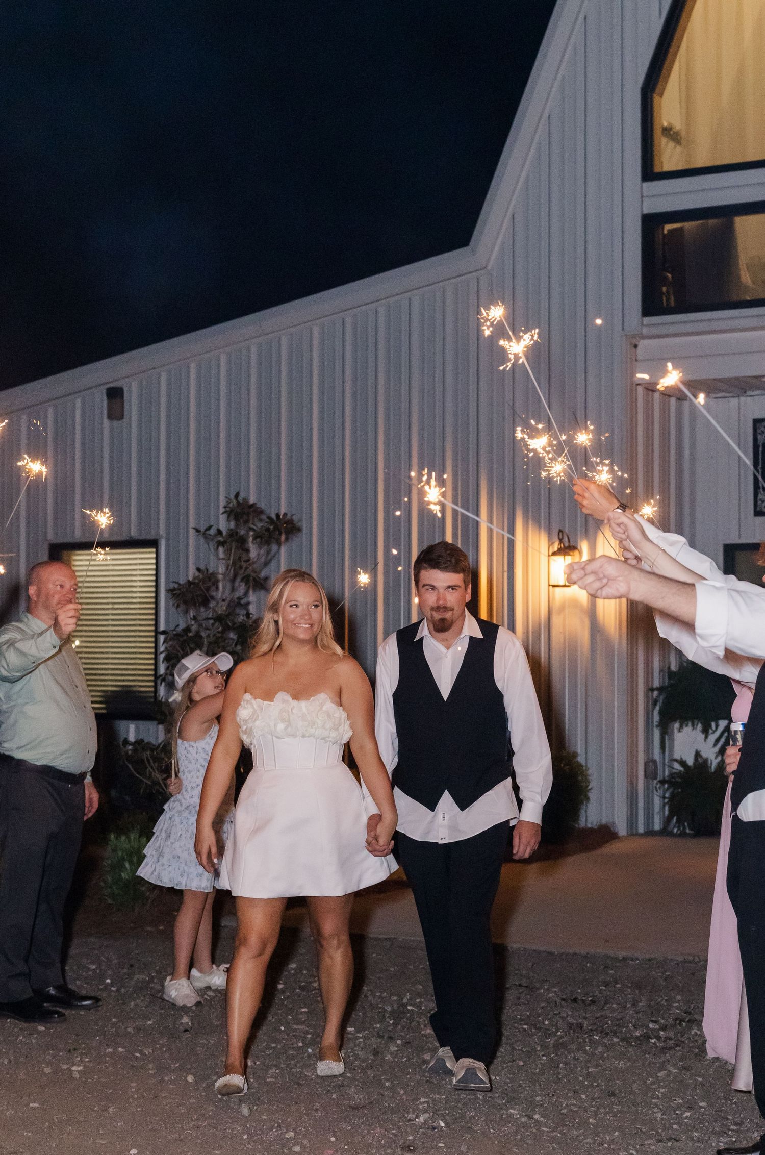 Bride and groom exit wedding venue with sparklers at night, guests celebrating around them.