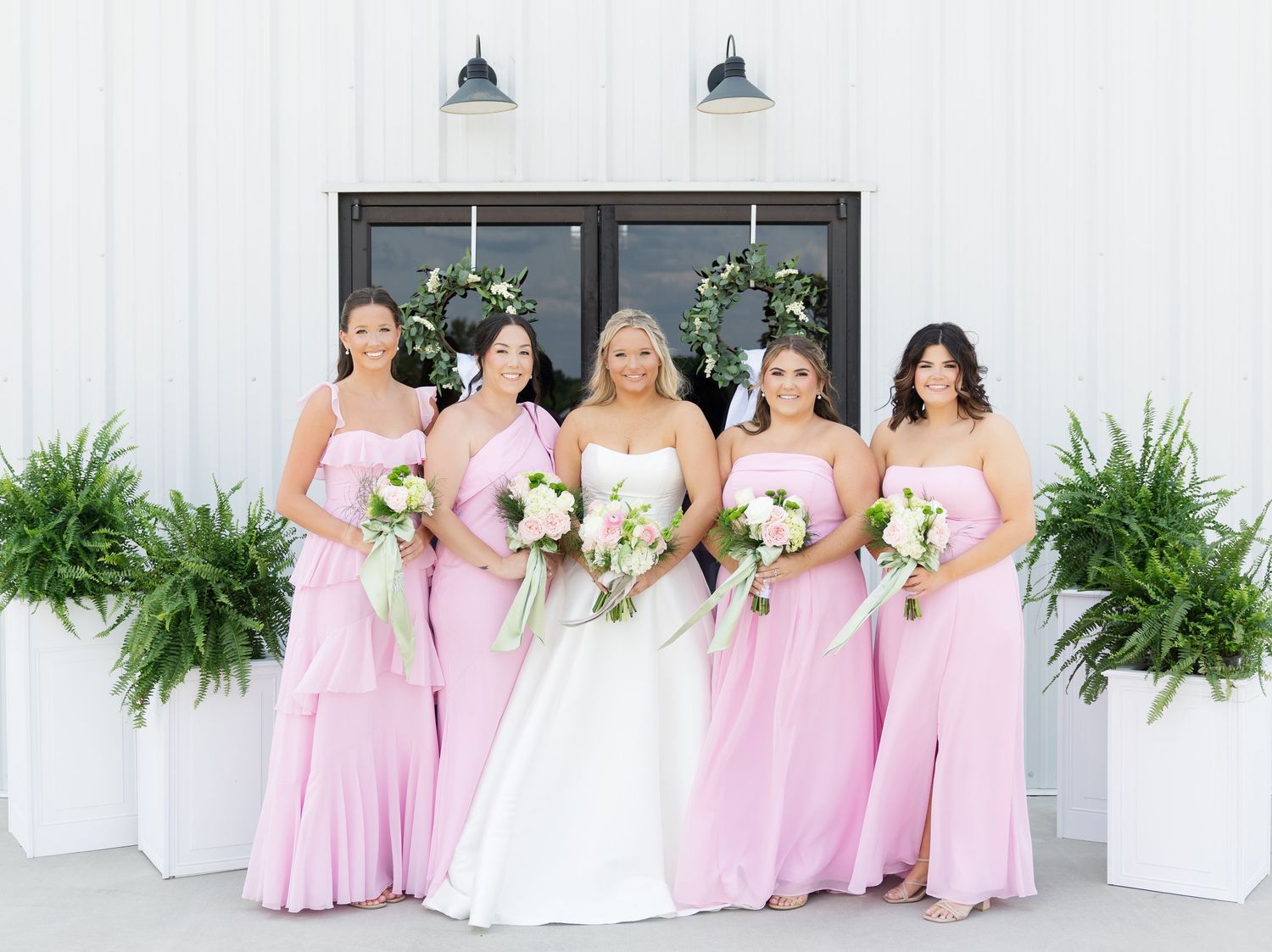 Bride in white gown with four bridesmaids in pink dresses holding bouquets outside a white barn venue.