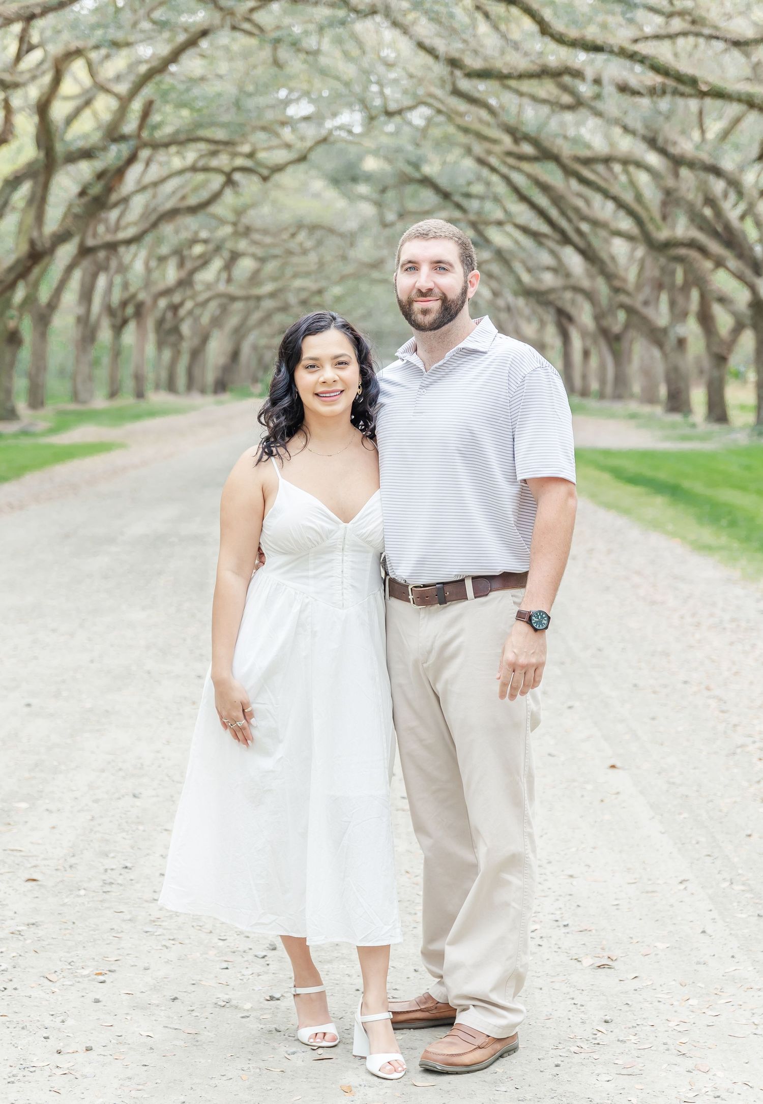 Couple in white and khaki clothing pose together on scenic tree-lined path.
