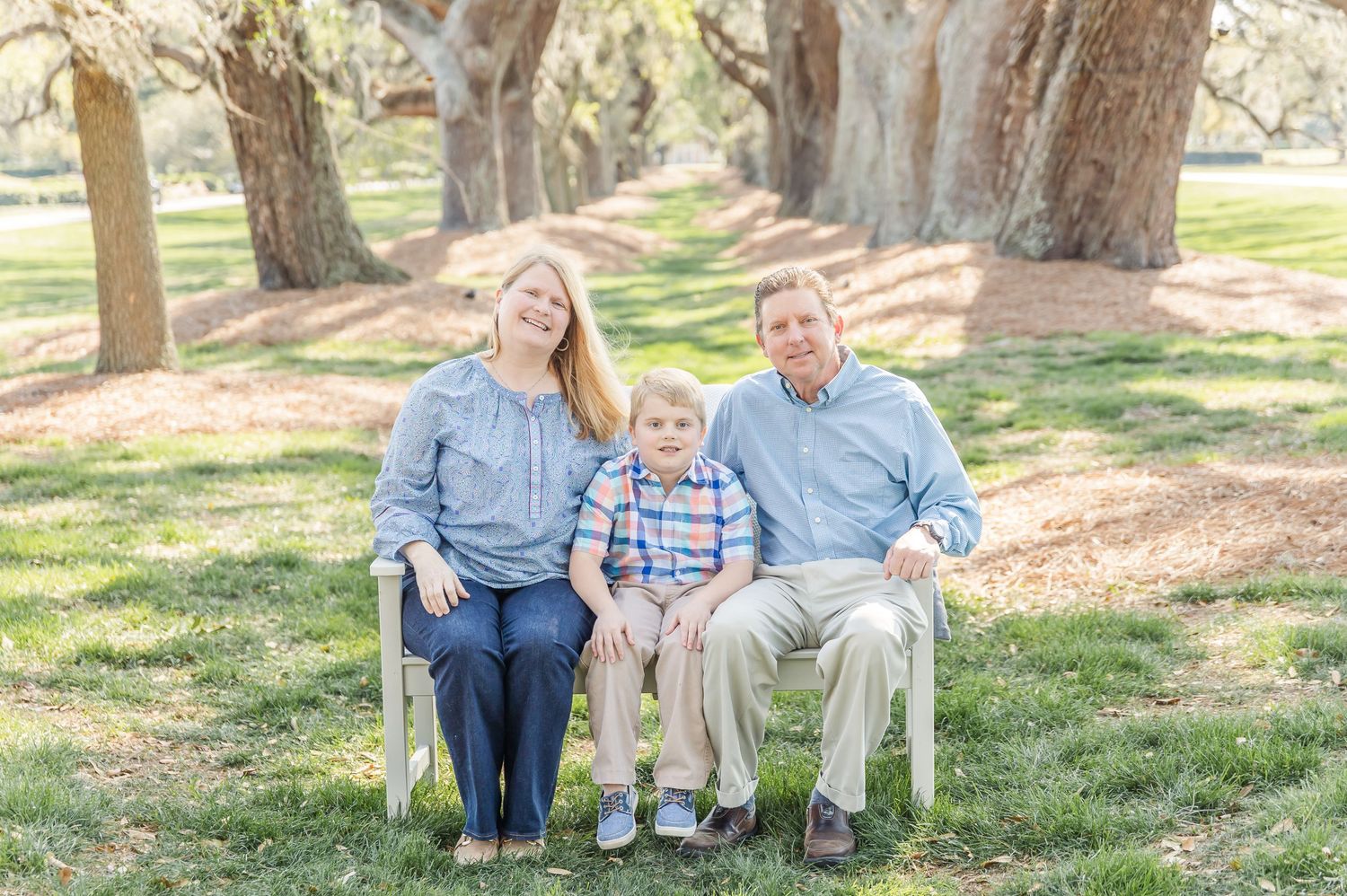 Family of three sitting together on white bench in sunlit oak tree grove.