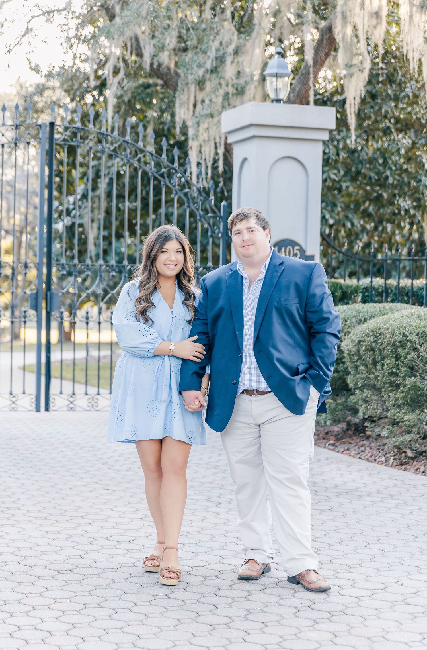 A couple poses together outdoors near a decorative iron gate, wearing coordinated blue and white outfits.