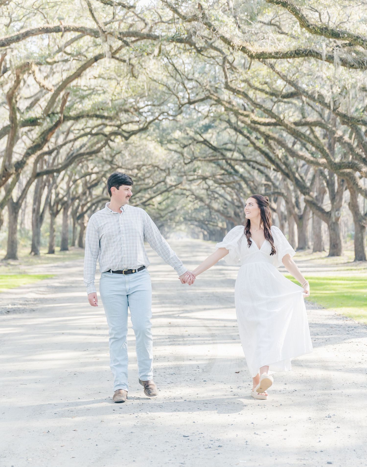 A couple skip together down a stunning tree-lined avenue in matching light outfits.