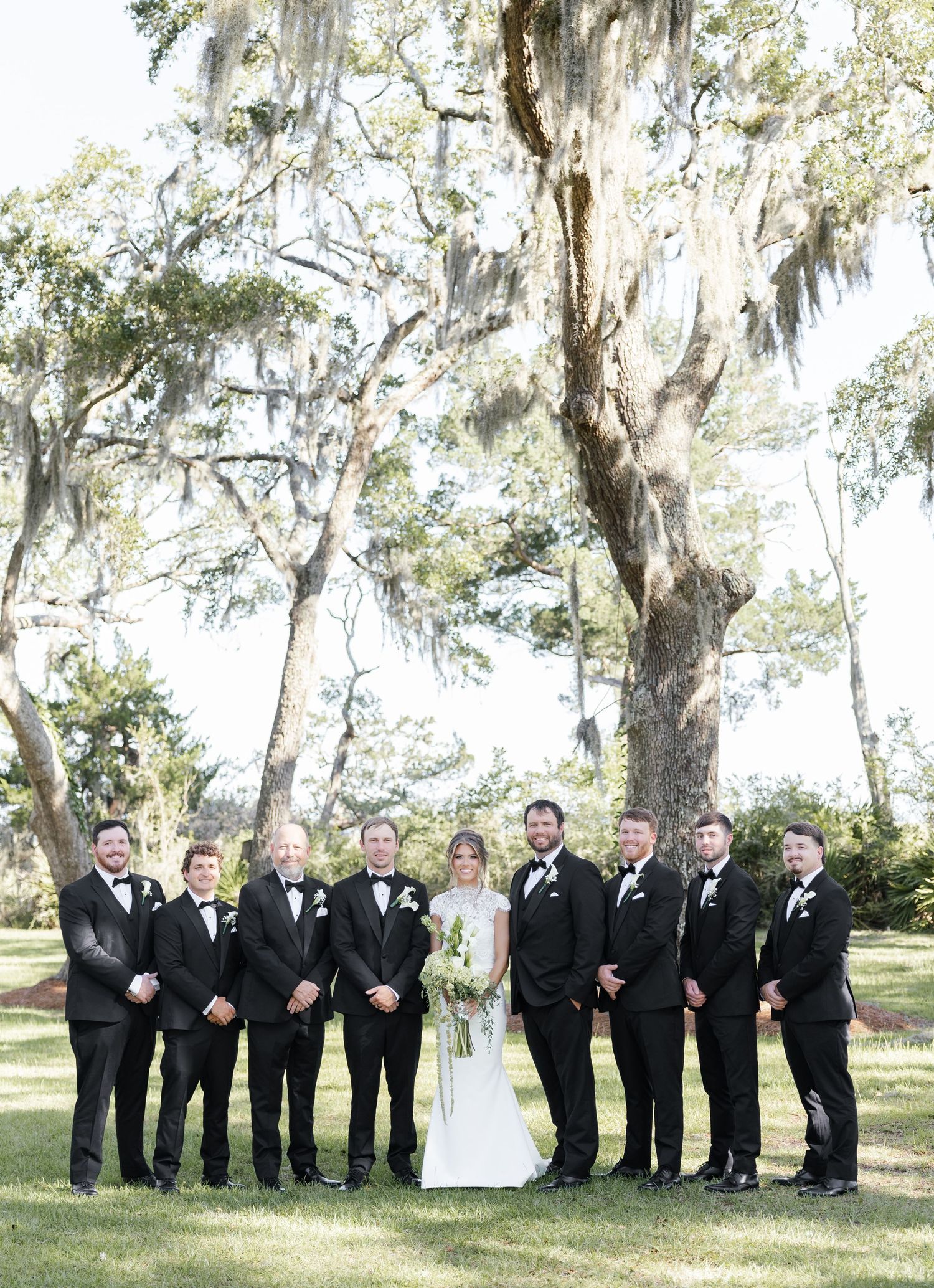 Wedding party poses outdoors under Spanish moss-draped oak trees, bride in white gown surrounded by groomsmen in black tuxedos.