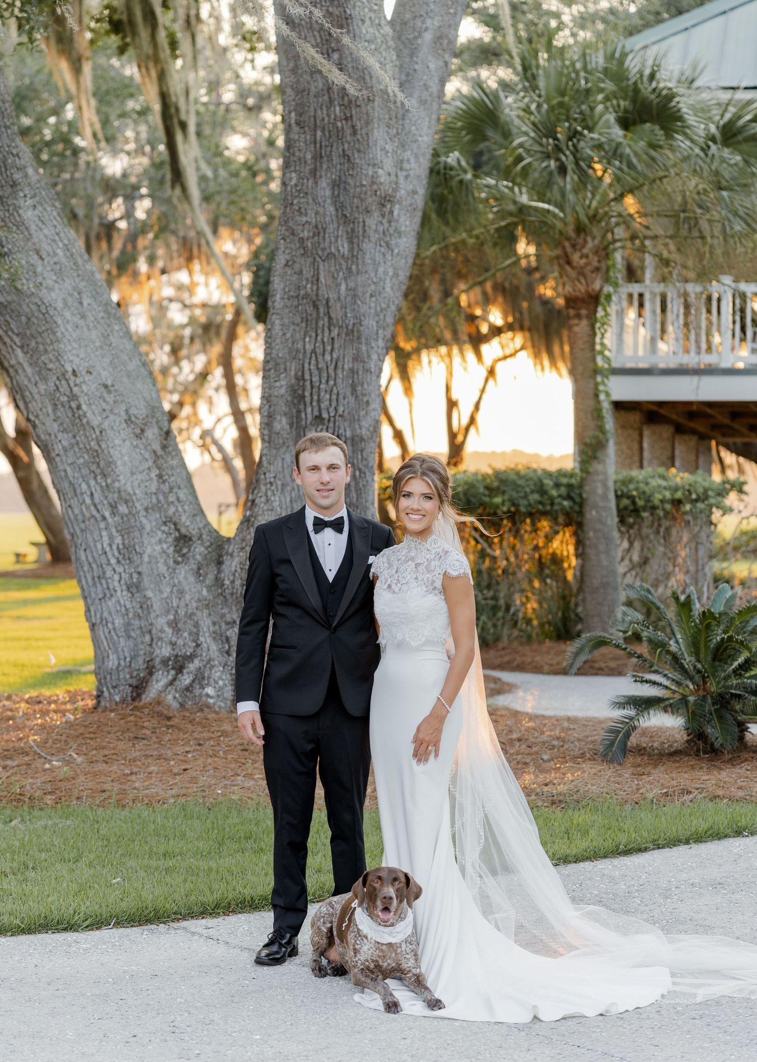 Bride and groom pose with their spotted dog outdoors near a large oak tree at a Southern wedding venue.