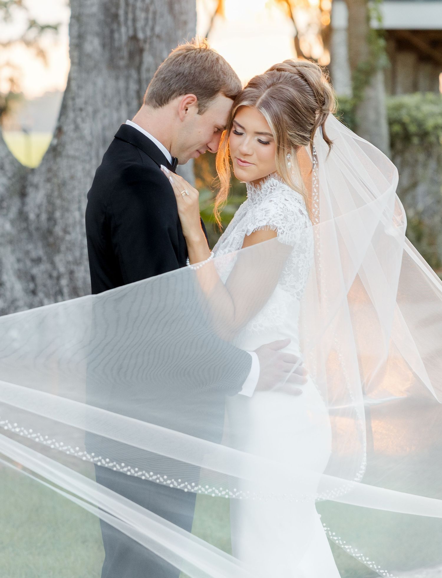 A bride in a lace gown and flowing veil leans close to her groom in a black tuxedo during a romantic outdoor portrait.
