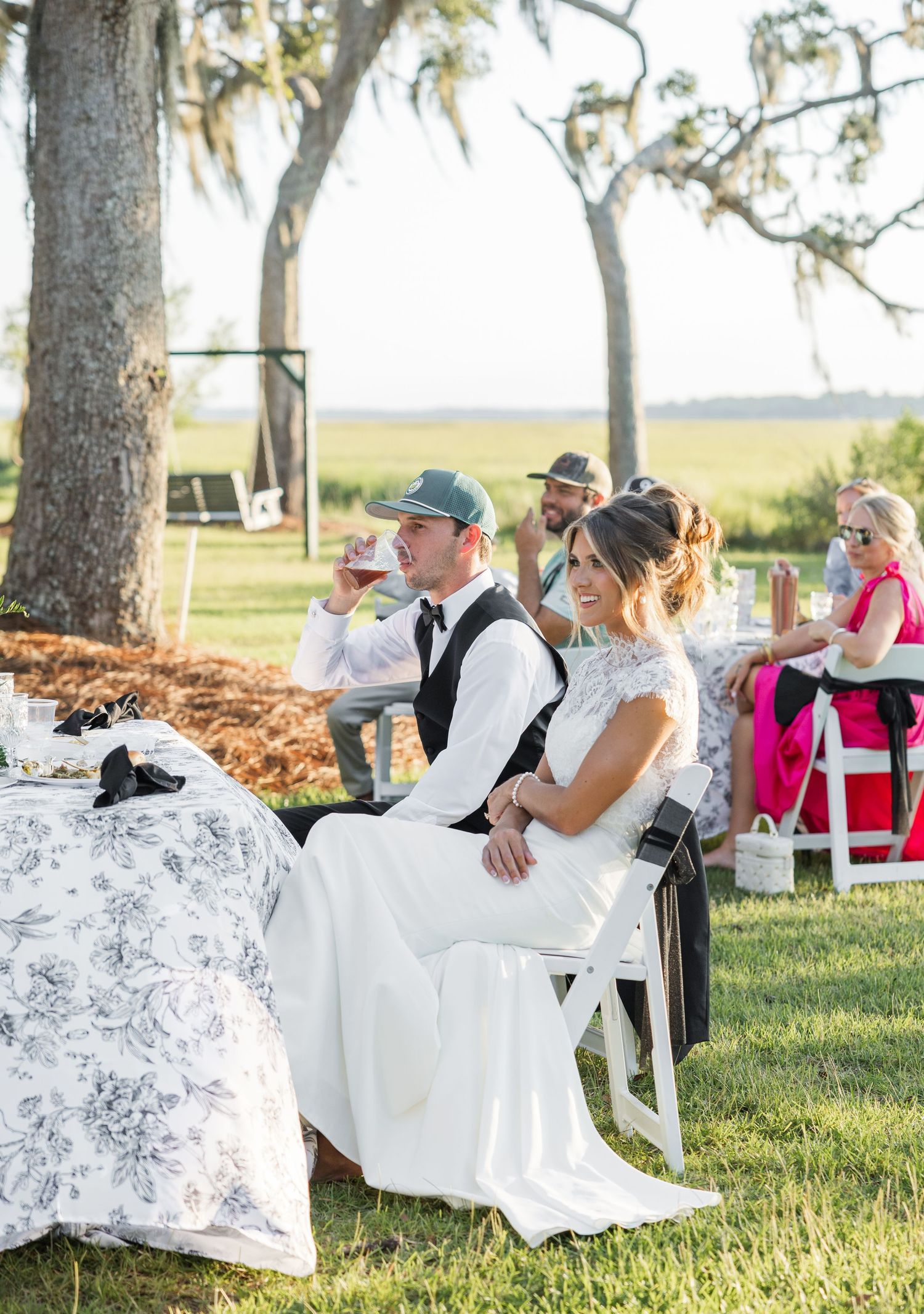 Bride and groom seated outdoors at elegant wedding reception with guests, trees, and scenic marsh views in background.