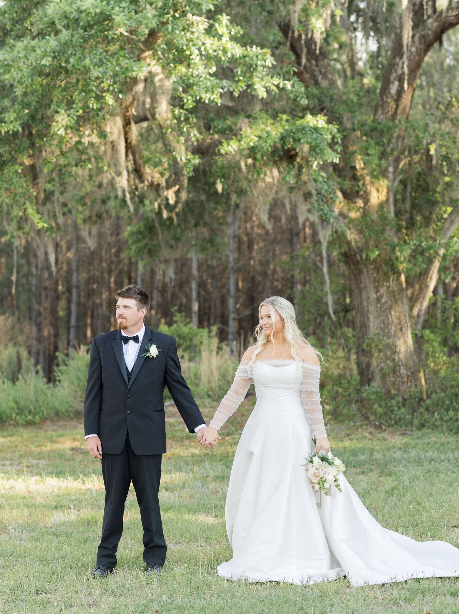 Bride and groom holding hands in a lush green field, she in a long-sleeve white gown, he in a black tuxedo.