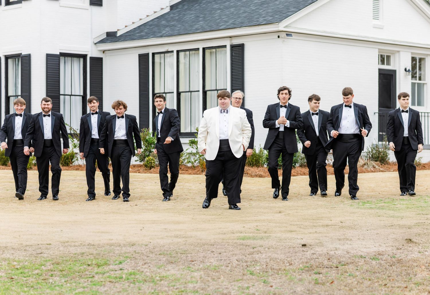 Groom in white jacket leads groomsmen in black tuxedos walking confidently outside a white plantation-style venue.