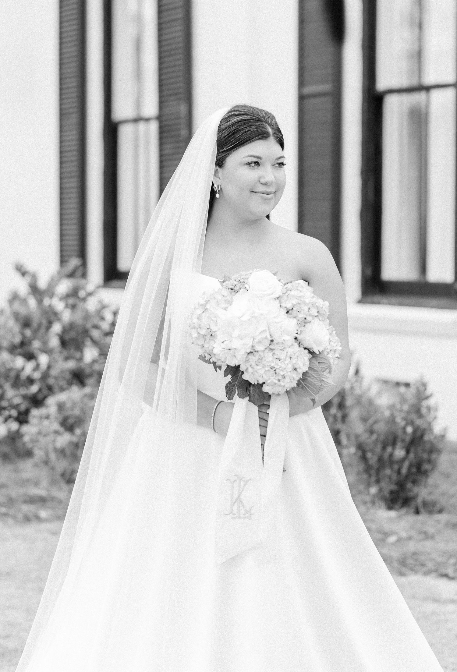 Black and white portrait of bride in strapless gown holding hydrangea bouquet with monogram ribbon.