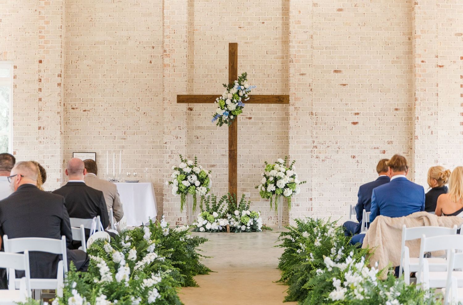 Wedding ceremony venue with wooden cross altar, white floral arrangements, and greenery aisle decor.