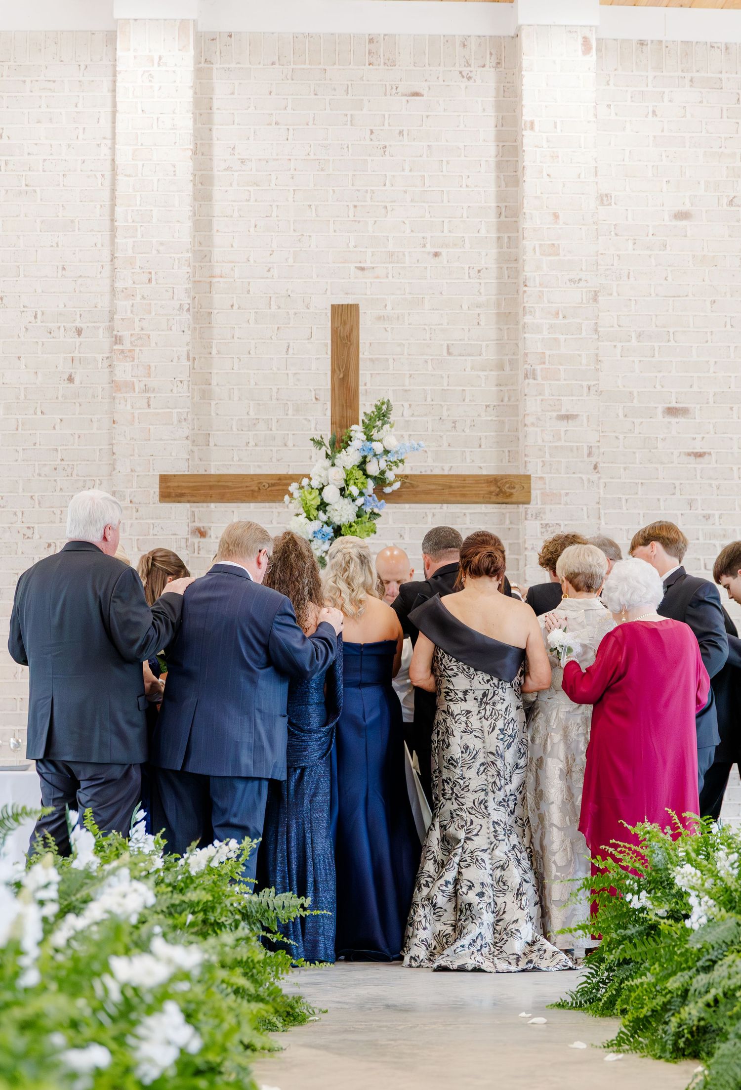 Wedding guests gather around couple for a prayer circle at the altar of a modern church with wooden cross.