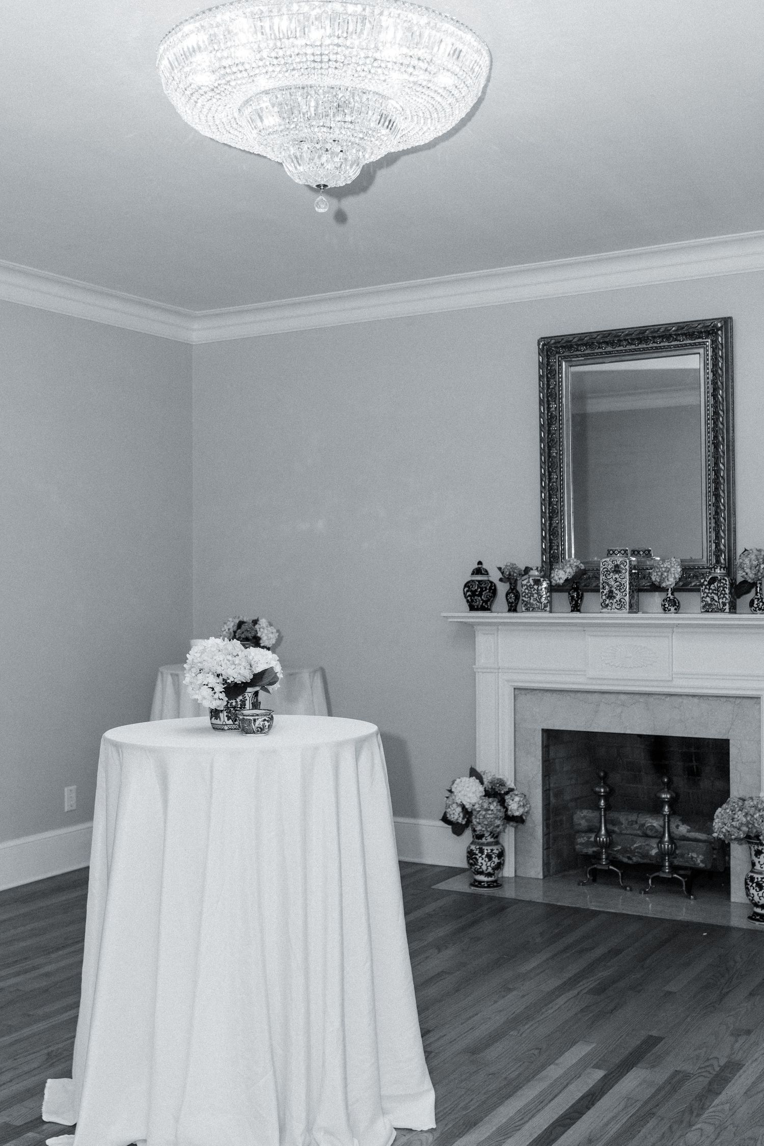 Black and white photo of elegant event space with crystal chandelier, fireplace, and cocktail table with floral arrangement.