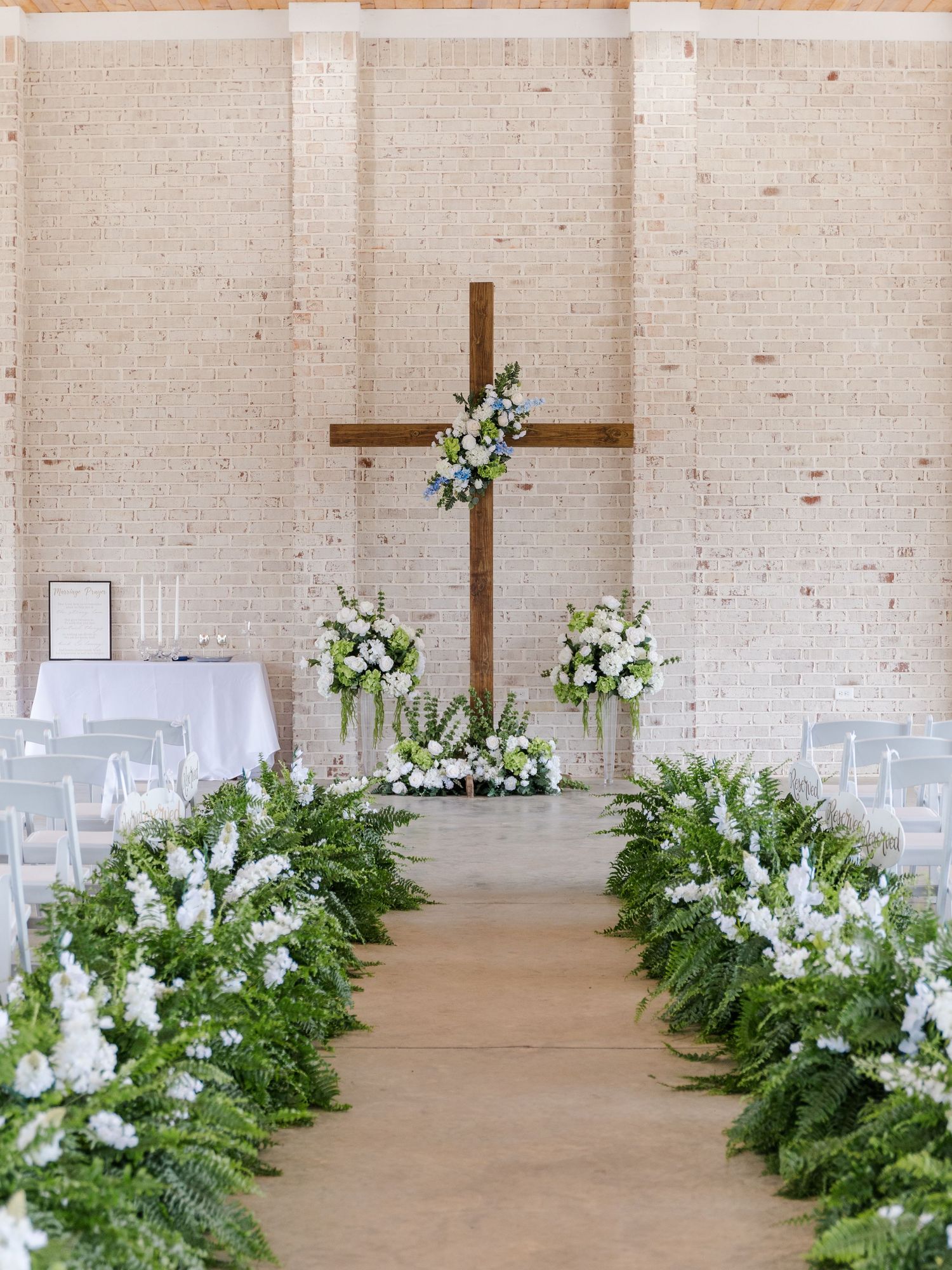 Church wedding ceremony aisle lined with lush ferns and white flowers leading to a wooden cross.