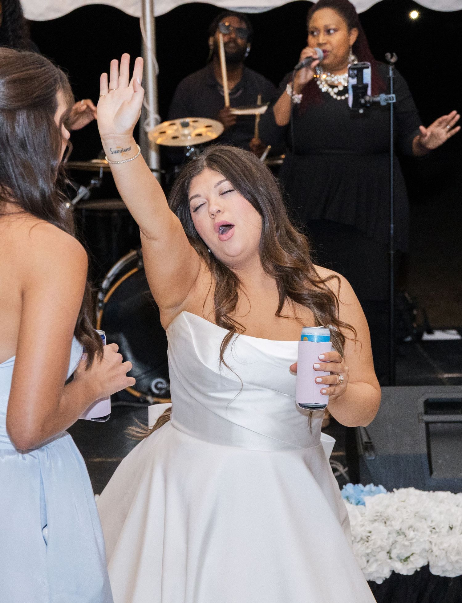 Joyful bride in strapless white gown high-fives bridesmaid while dancing at wedding reception with live band performing.