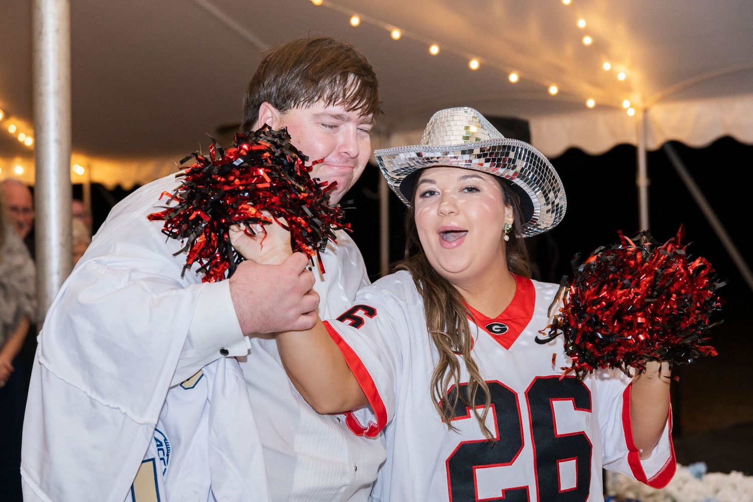 Bride and groom in football jerseys celebrating with red pom-poms and silver cowboy hat at reception.