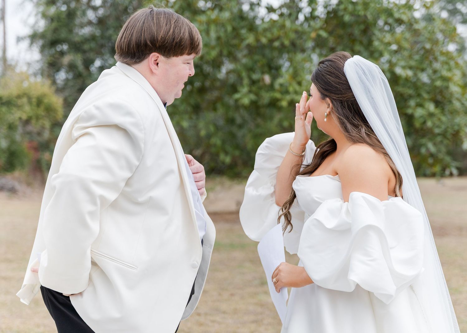 Emotional bride wipes tears during touching first look reveal with groom in white tuxedo jacket outdoors.