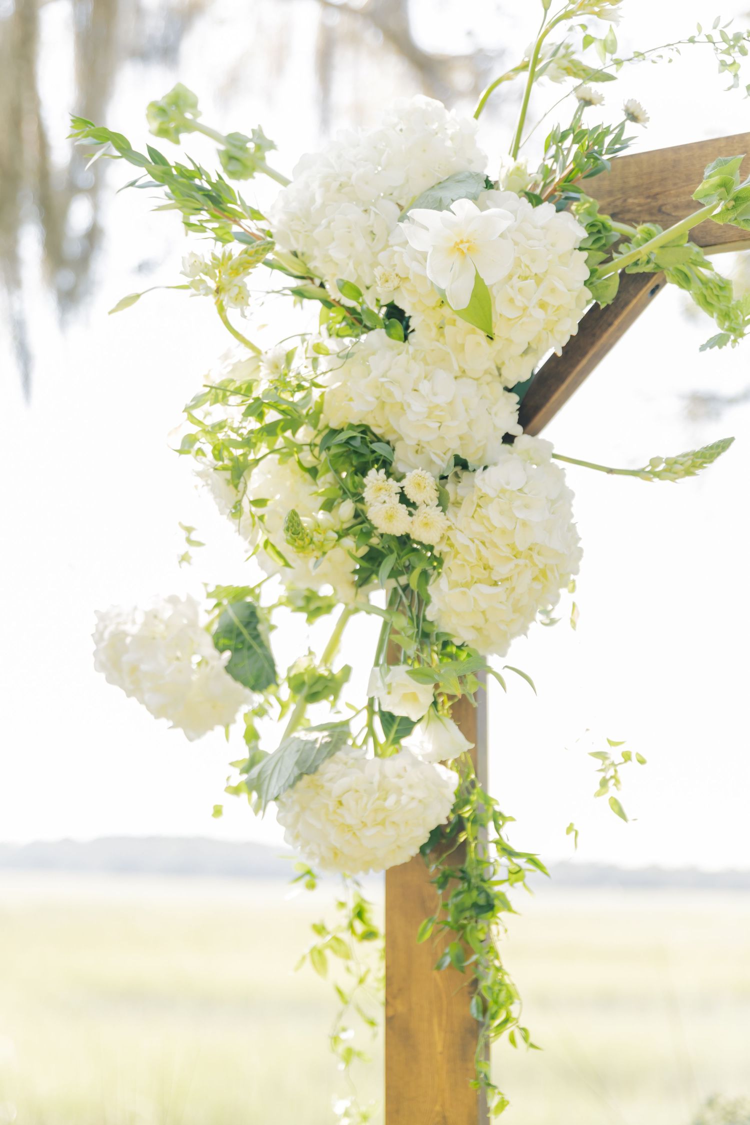 White hydrangea and greenery floral arrangements decorating wooden wedding arch posts.
