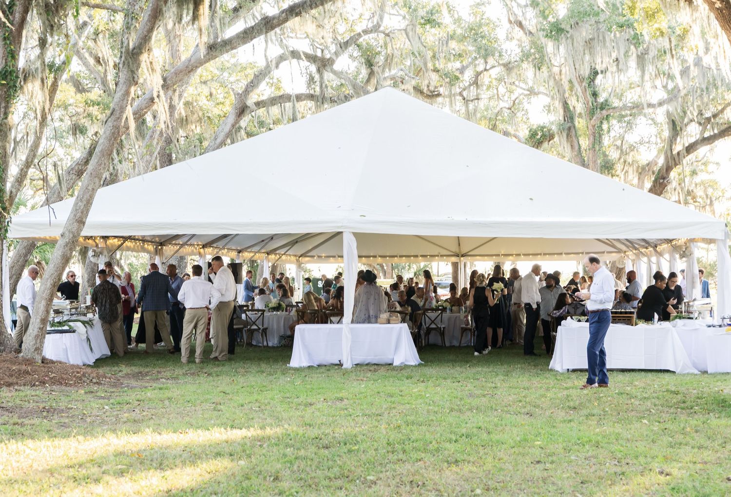 Large white tent set up for outdoor wedding reception under oak trees.