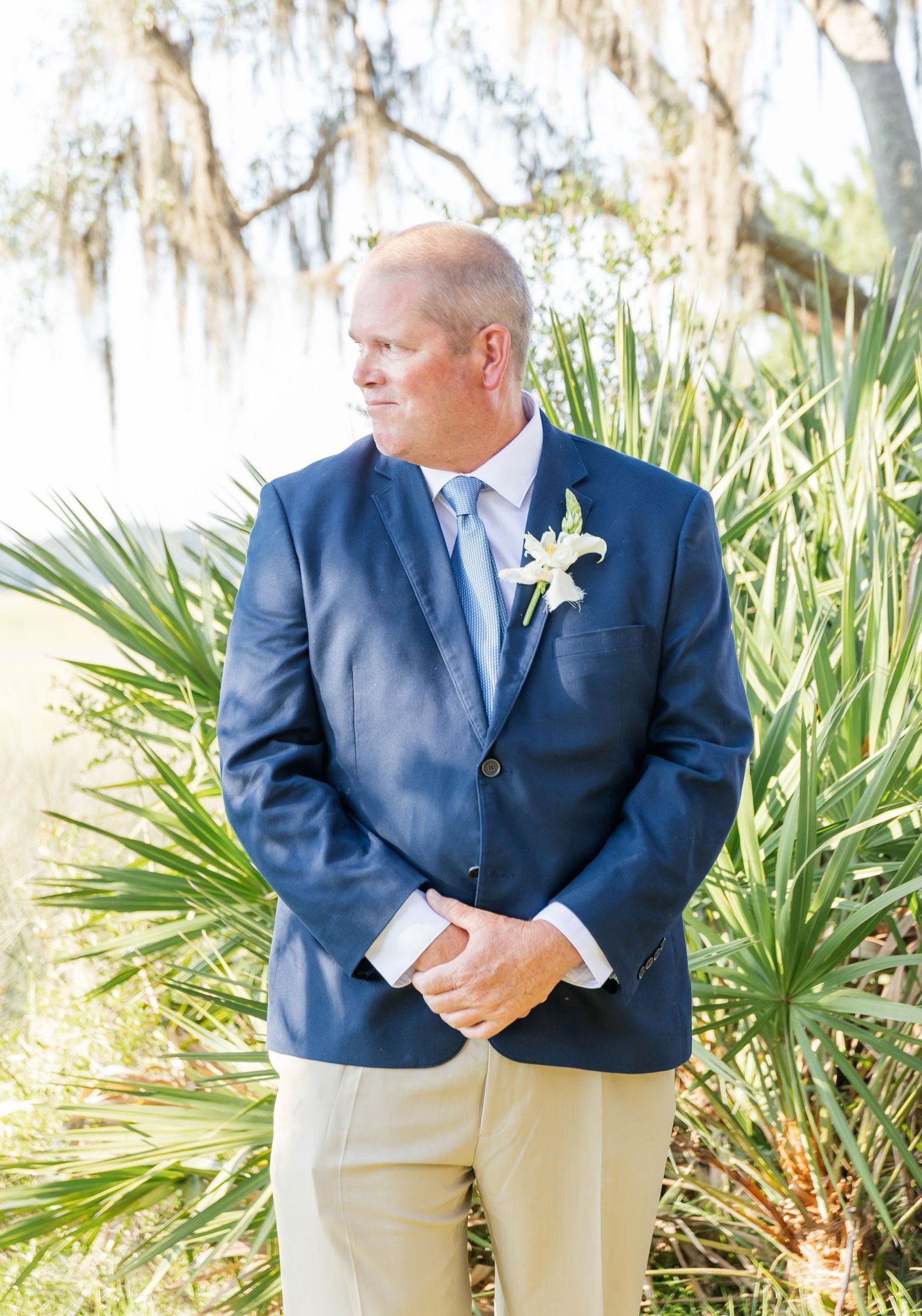 A person in a navy blue suit jacket with a white boutonniere stands against a backdrop of palm fronds and Spanish moss.