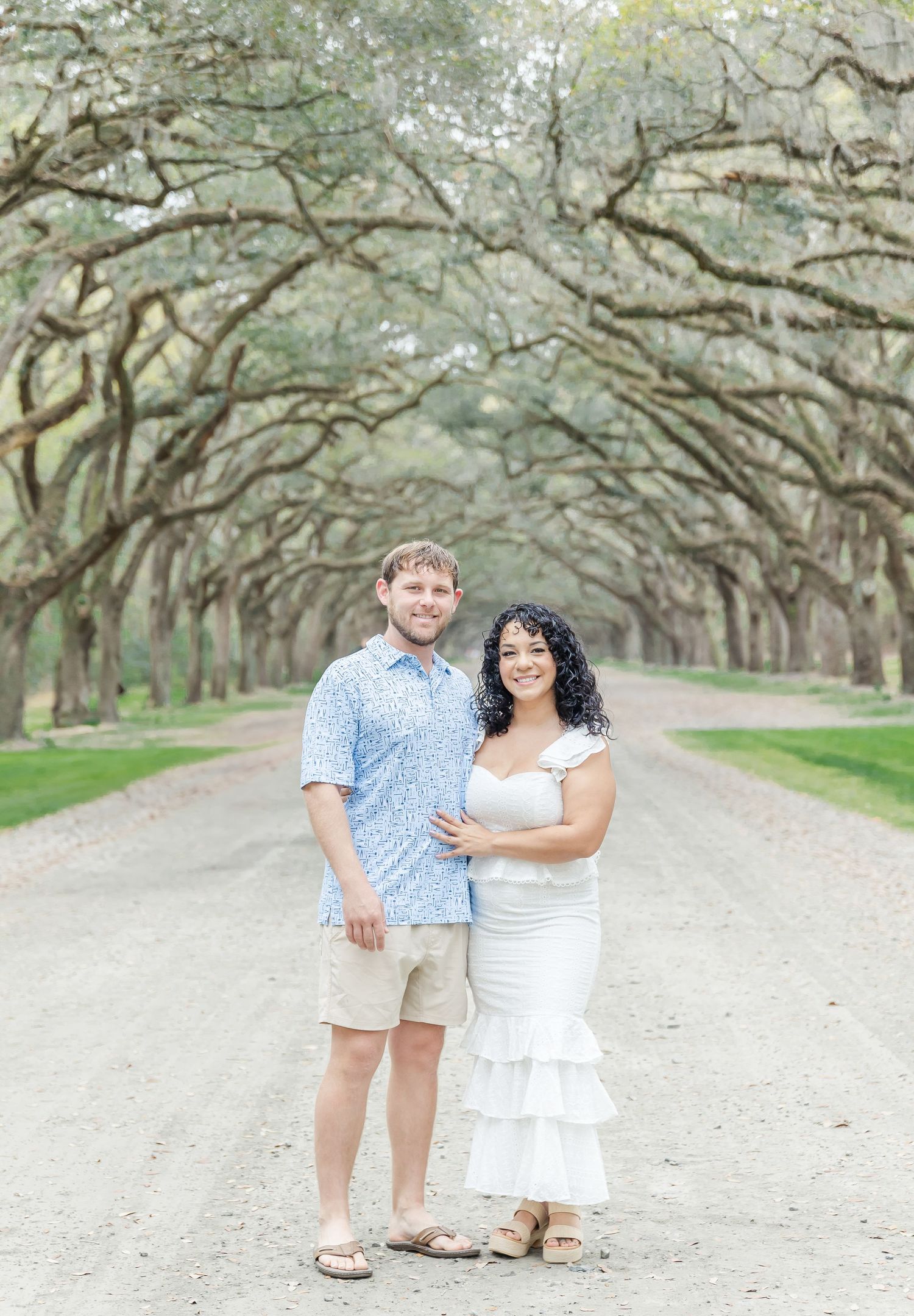 Couple in casual attire standing together on a scenic tree-lined path.