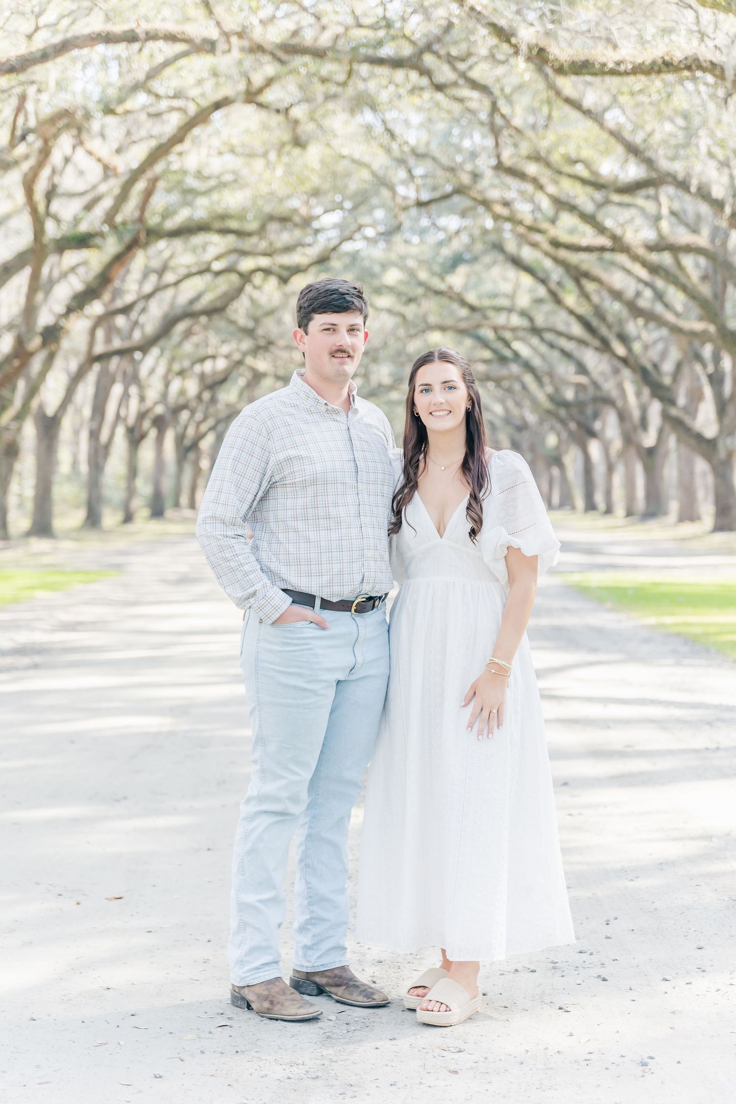 A couple stands together on a dreamy oak-lined path in light spring attire.