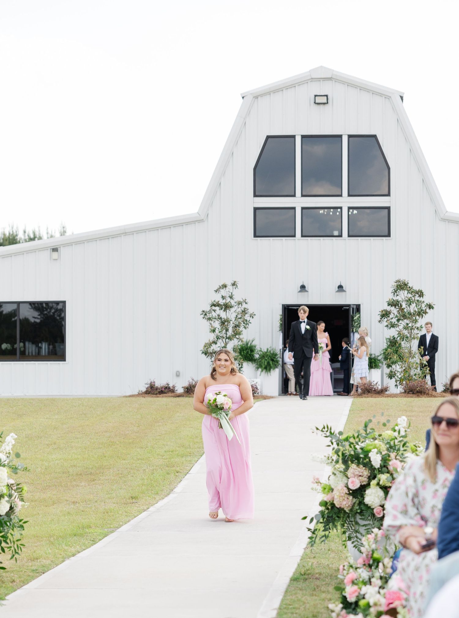 Bridesmaid in pink gown walking down the outdoor aisle toward guests at a white barn wedding venue.