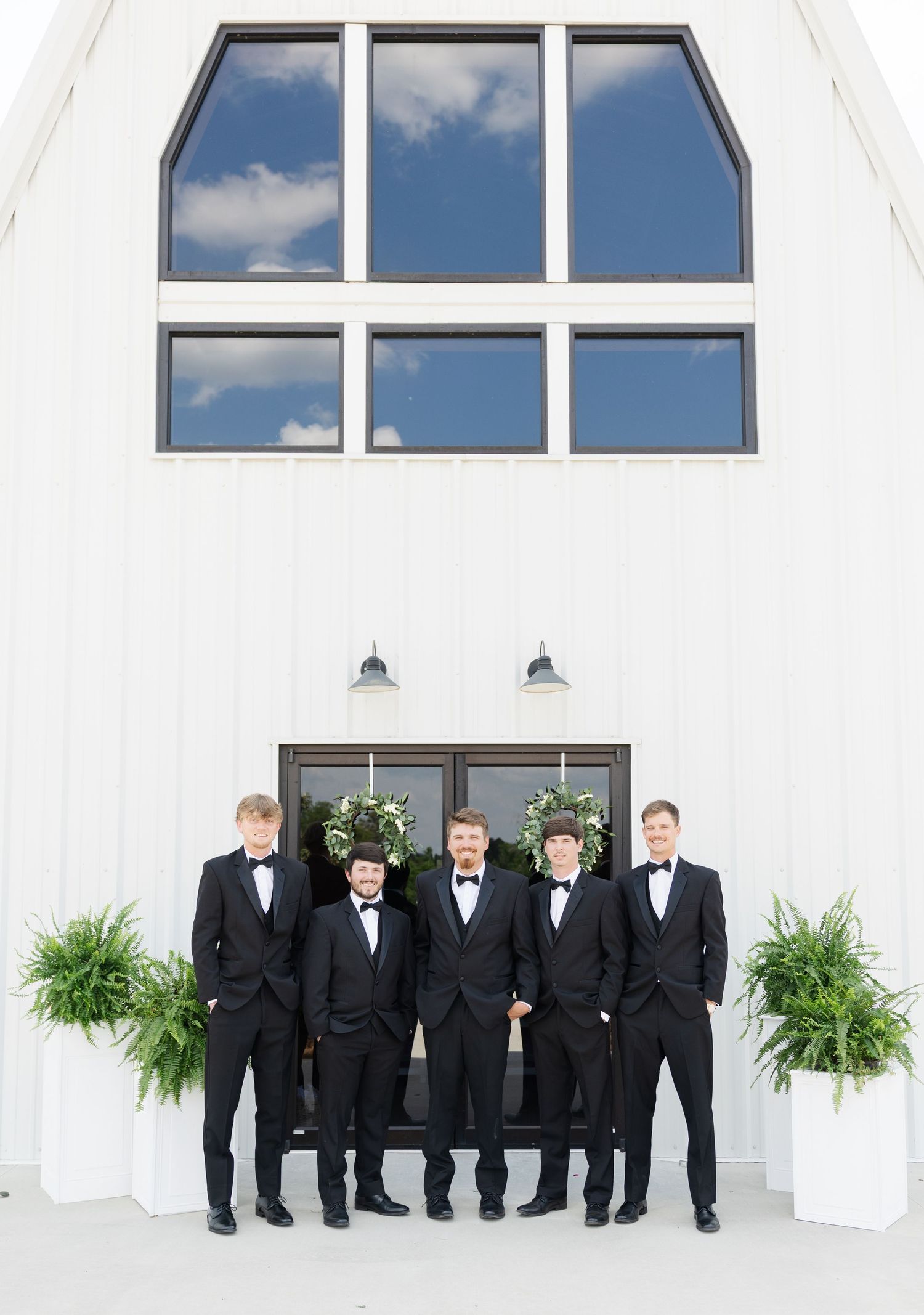 Five groomsmen in black tuxedos stand before a white barn wedding venue with large arched windows and lush greenery.