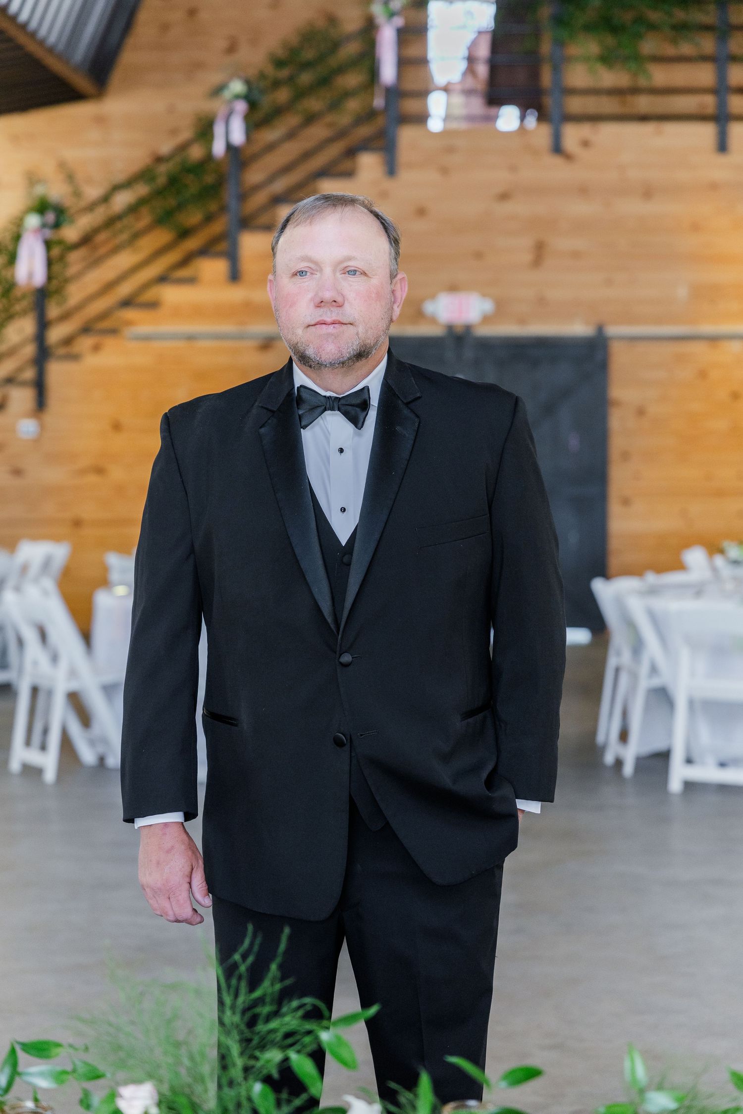 Groom in black tuxedo with bow tie stands in rustic wood-paneled wedding venue with white chairs.